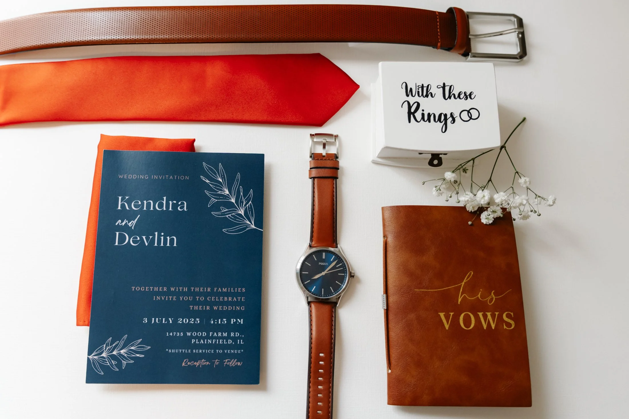 Wedding invitation with the names Kendra and Devlin, date and time of the wedding, wedding details, a brown leather strap watch, a brown vow book labeled 'his VOWS', a white box with 'With these Rings' written on it, an orange and beige belt, an oran