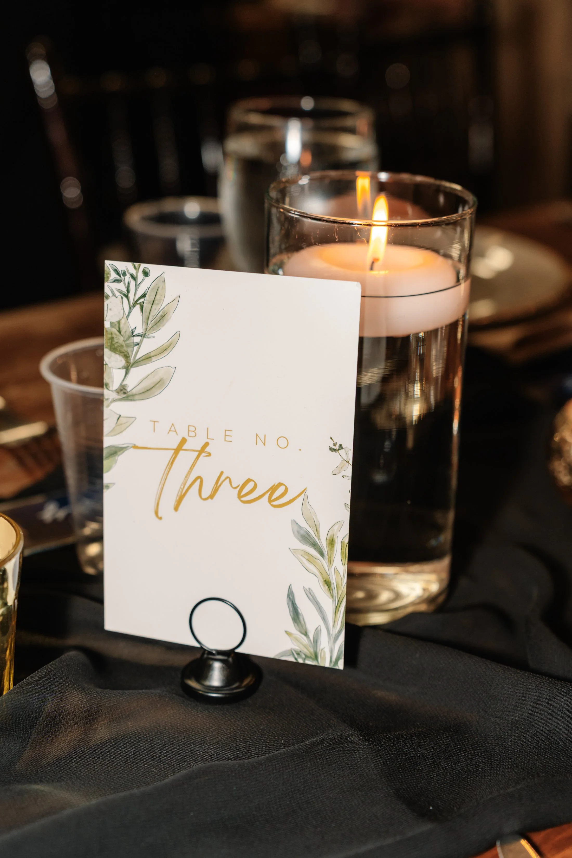 A table setting with a candle in a glass holder, a table number card labeled 'Table No. Three' with botanical illustrations, and glasses in the background.