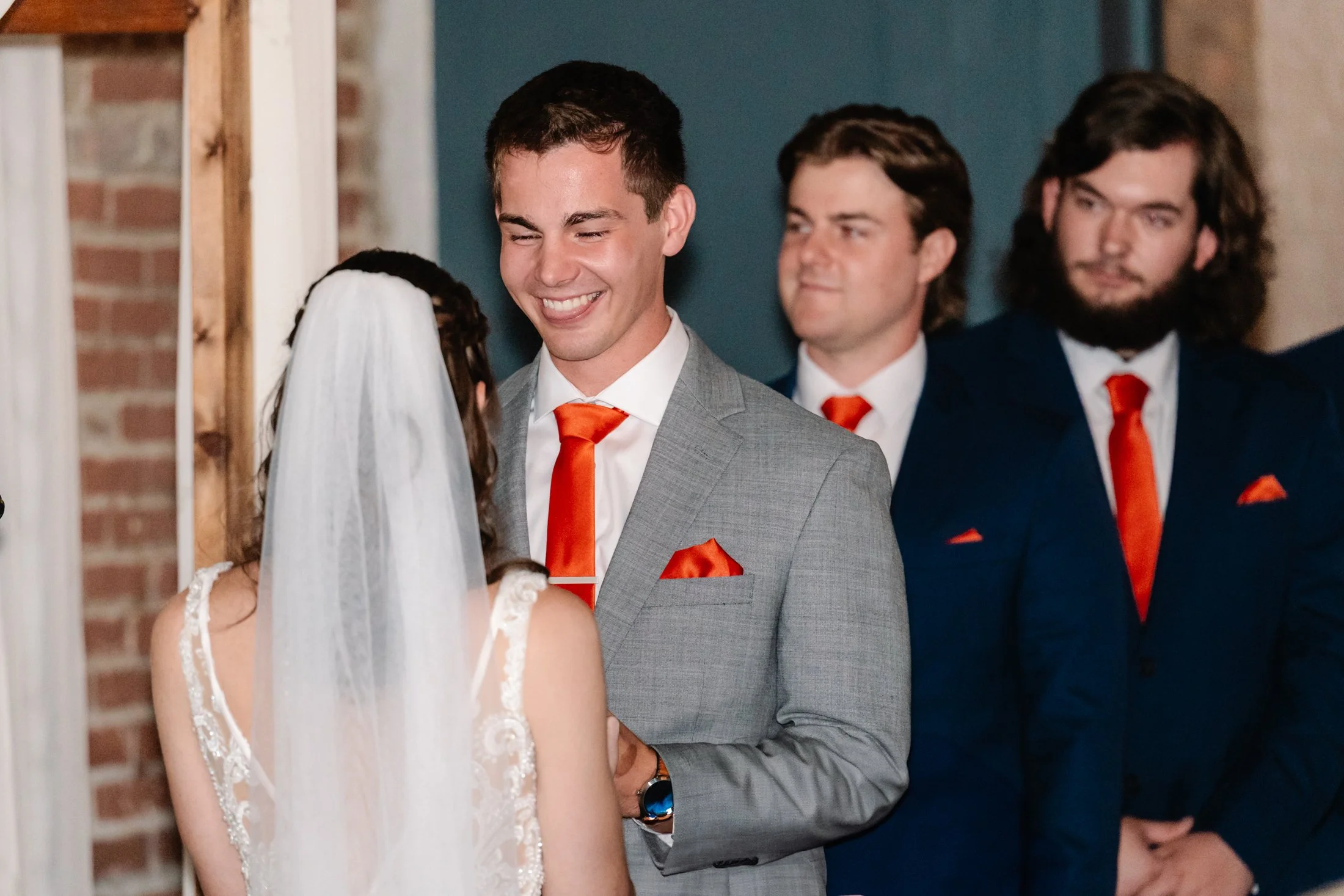 A groom in a gray suit and orange tie smiling at a bride during their wedding ceremony, with two groomsmen in blue suits and orange ties in the background.