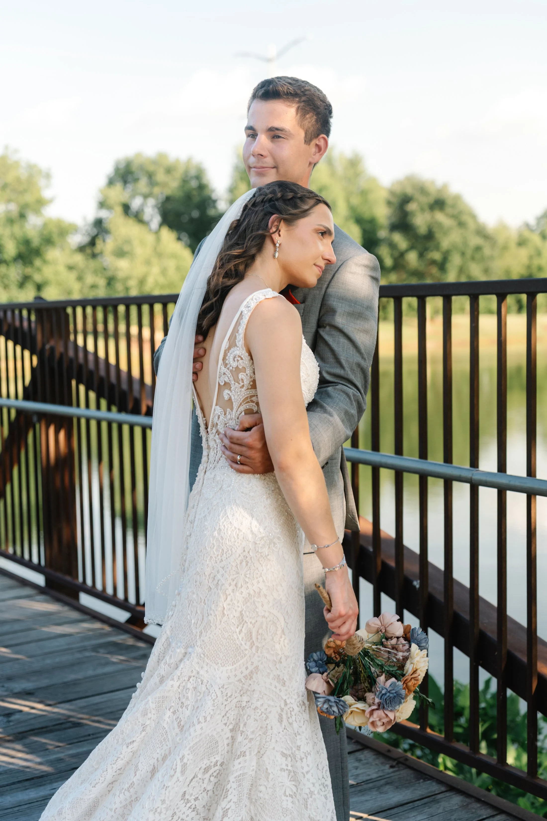 A bride and groom standing on a wooden bridge, embracing each other. The bride is holding a bouquet and wearing a lace wedding dress, and the groom is in a gray suit. Behind them, there is a scenic view of green trees and a body of water.