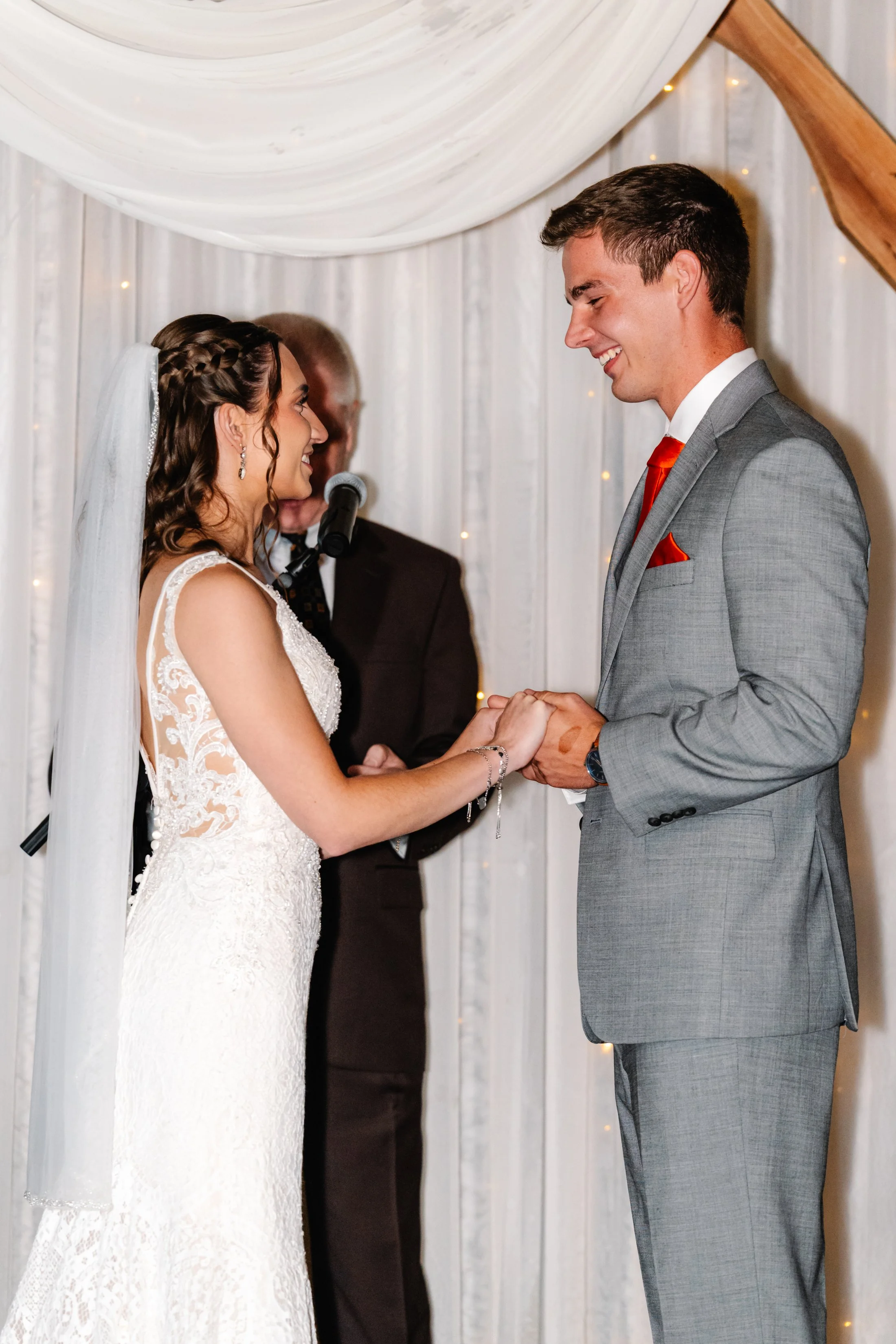 Bride and groom holding hands during their wedding vows, standing in front of officiant with white drapery and string lights.