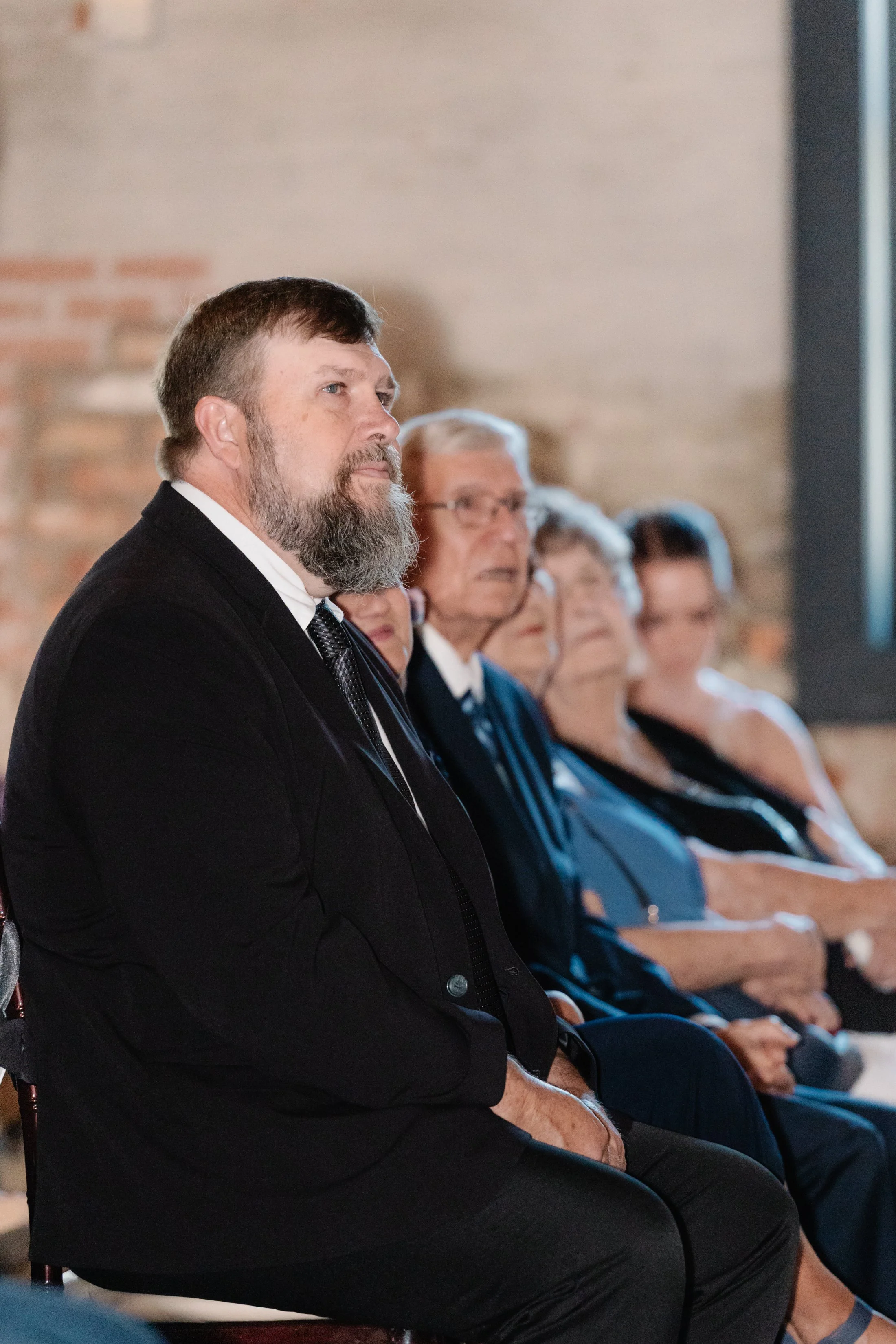 A group of people sitting in a row watching an event, with the focus on a bearded man in a black suit in the foreground.