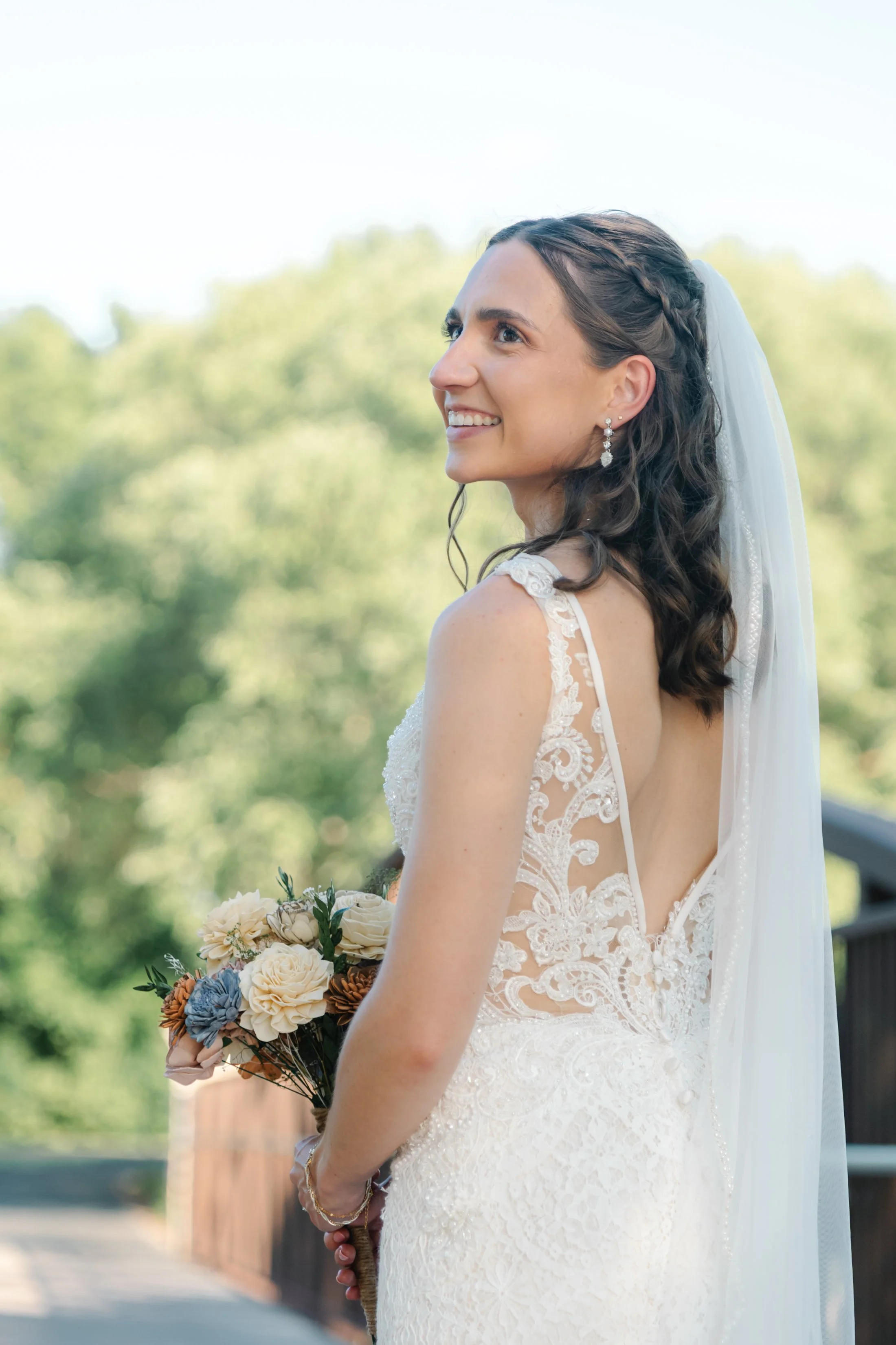 Bride in a lace wedding dress holding a bouquet, smiling outdoors.