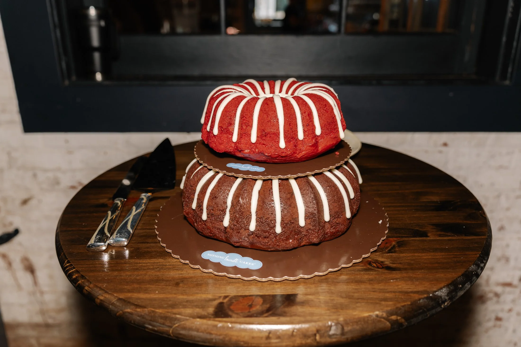 Two stacked bundt cakes, one red and one chocolate, decorated with white icing stripes, placed on a wooden table with cake serving utensils nearby.