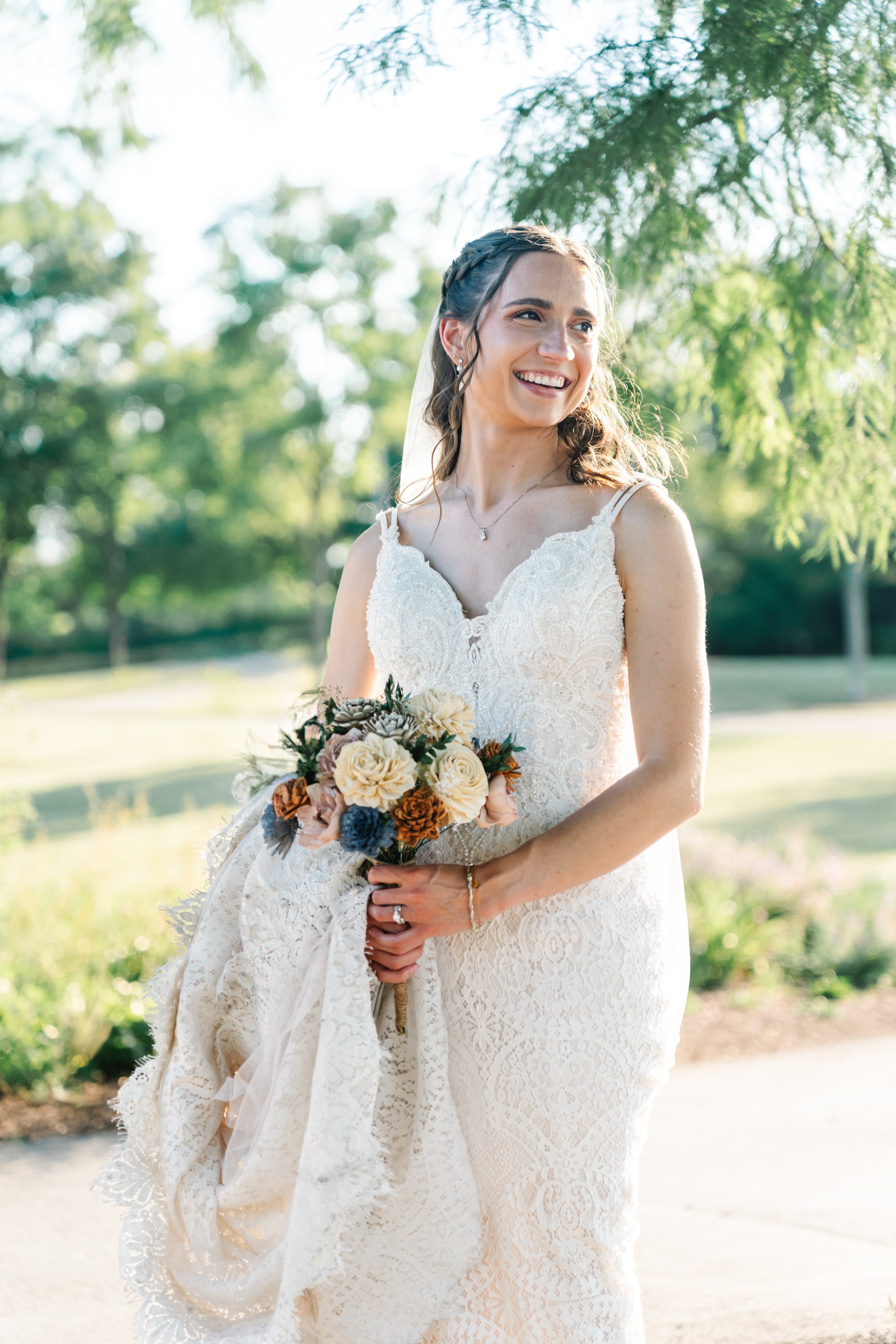 A smiling bride in a white lace wedding gown holding a bouquet of cream, brown, and blue flowers outdoors in a park setting with greenery and trees.