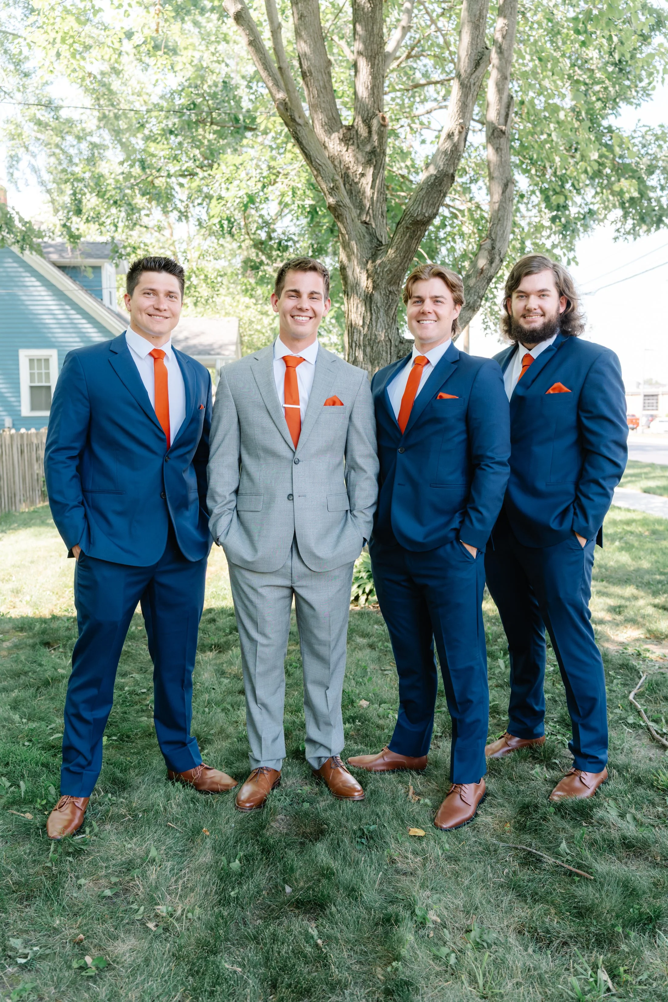 Four men in suits with orange ties and pocket squares standing outdoors in front of a tree and house, smiling for a group photo.