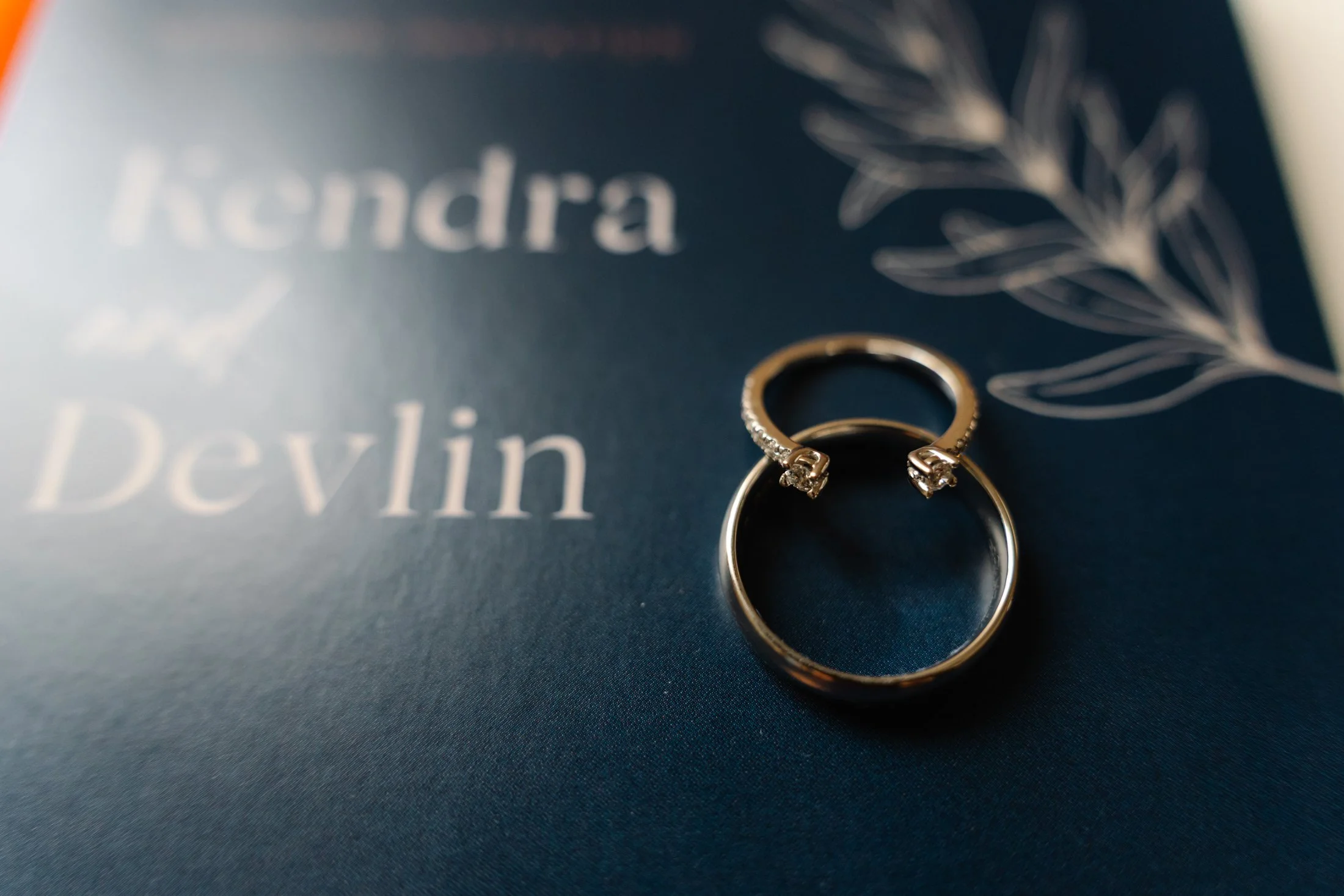 Close-up of two gold wedding rings with small diamonds on a dark blue textured background, with blurred white text and a white leaf outline.