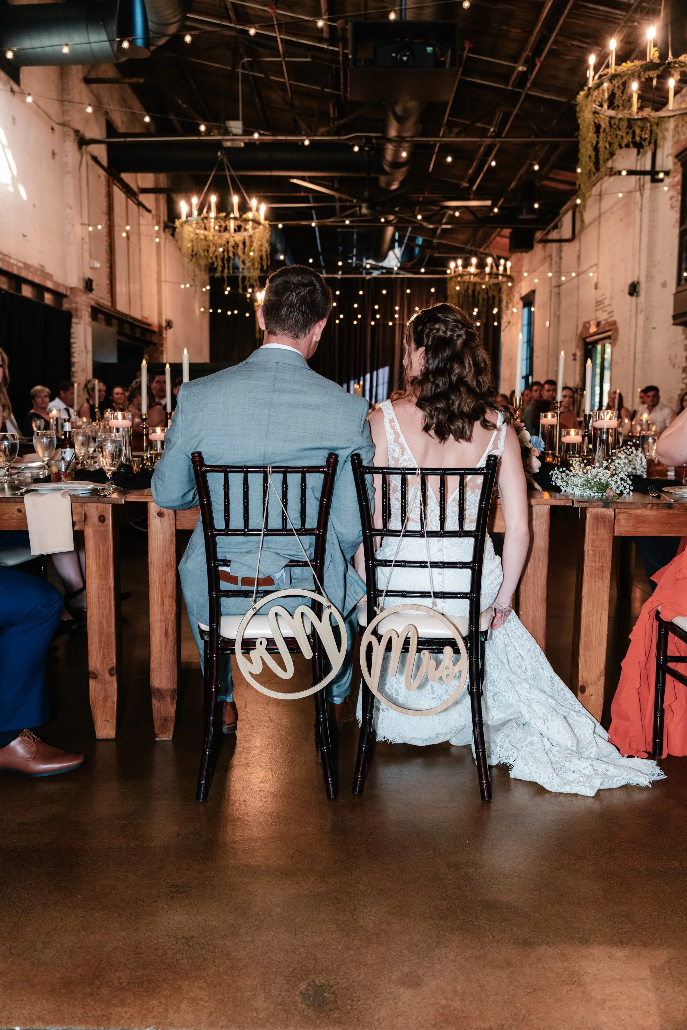 Bride and groom sitting at a wedding reception table with signed 'Mr' and 'Mrs' signs hanging on their chairs, decorated with candles and flowers, in an industrial-style venue.
