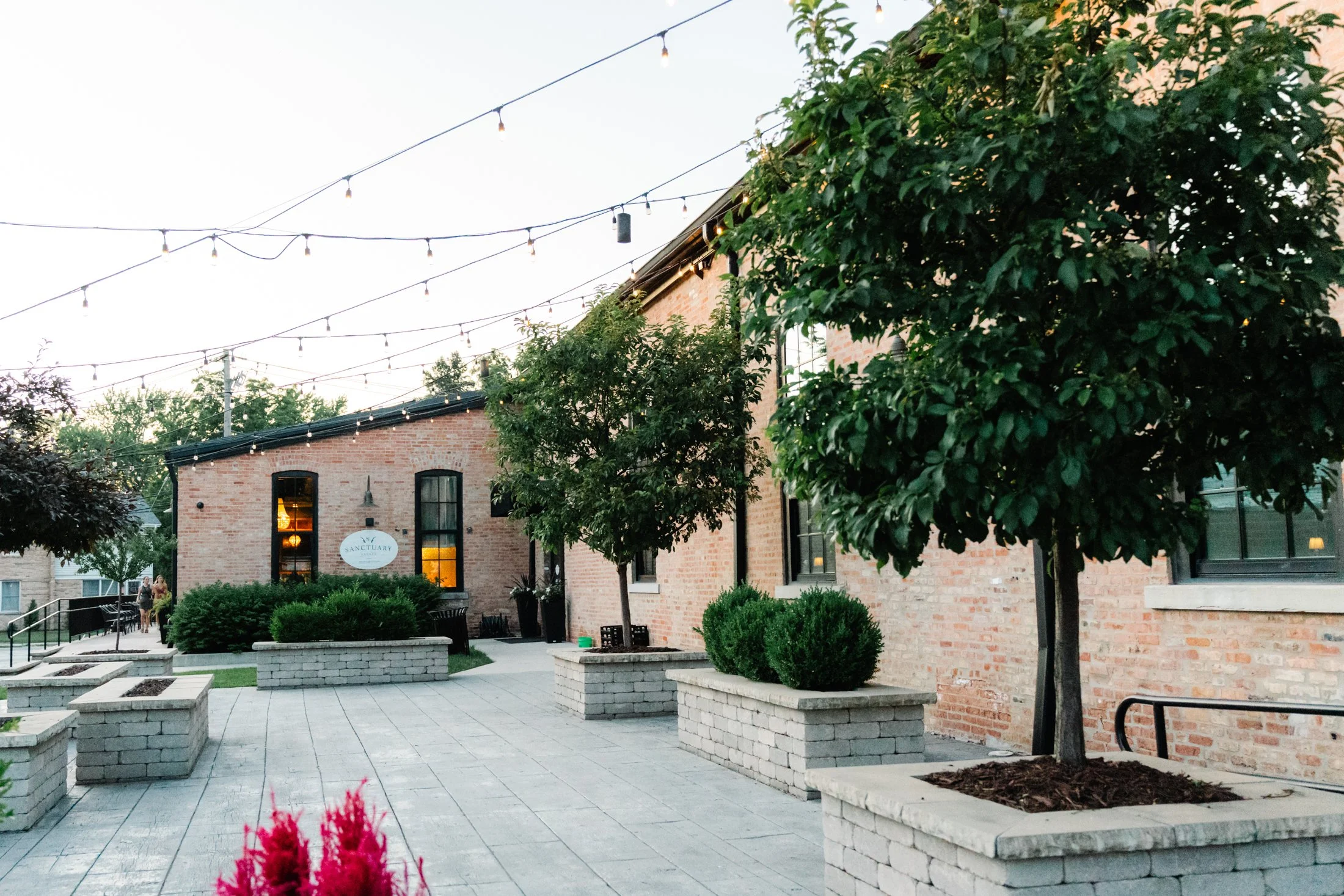 Outdoor patio area with brick building, string lights overhead, and trees in large brick planters, with some people walking in the background.