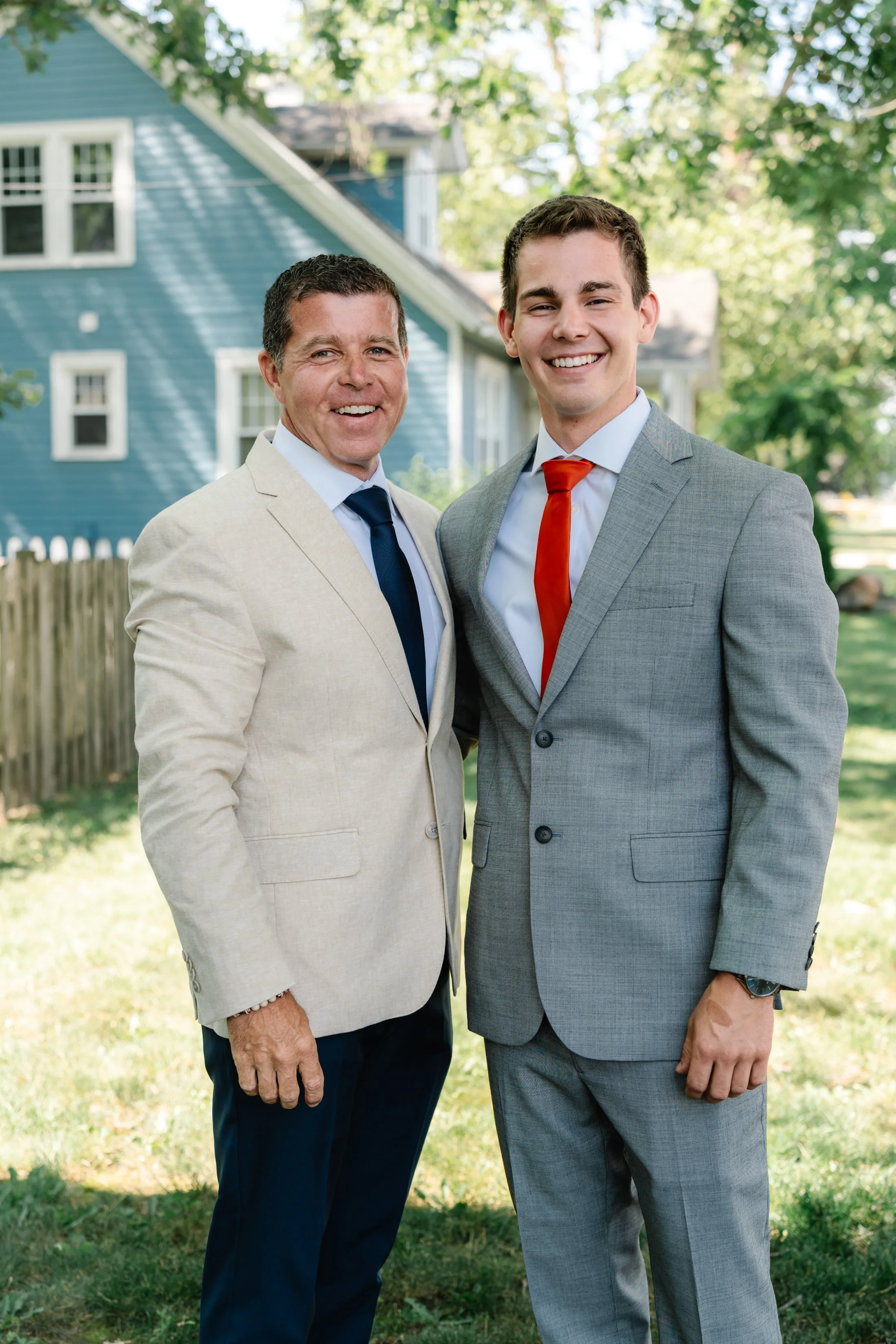 Two men in suits standing outdoors in front of a blue house with trees in the background, smiling at the camera.