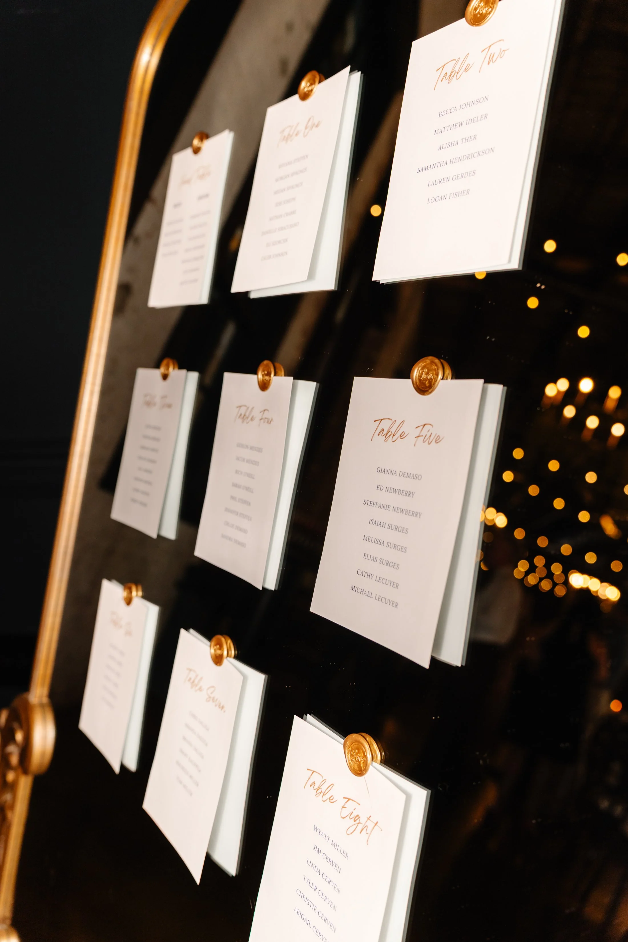 A wedding seating chart with white cards listing guest names for each table, displayed with gold push pins on a black mirror background.