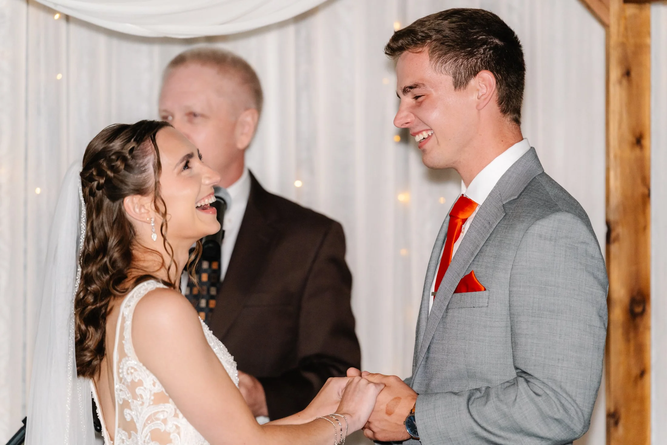 A bride and groom smiling and holding hands during their wedding ceremony, with an officiant in the background.
