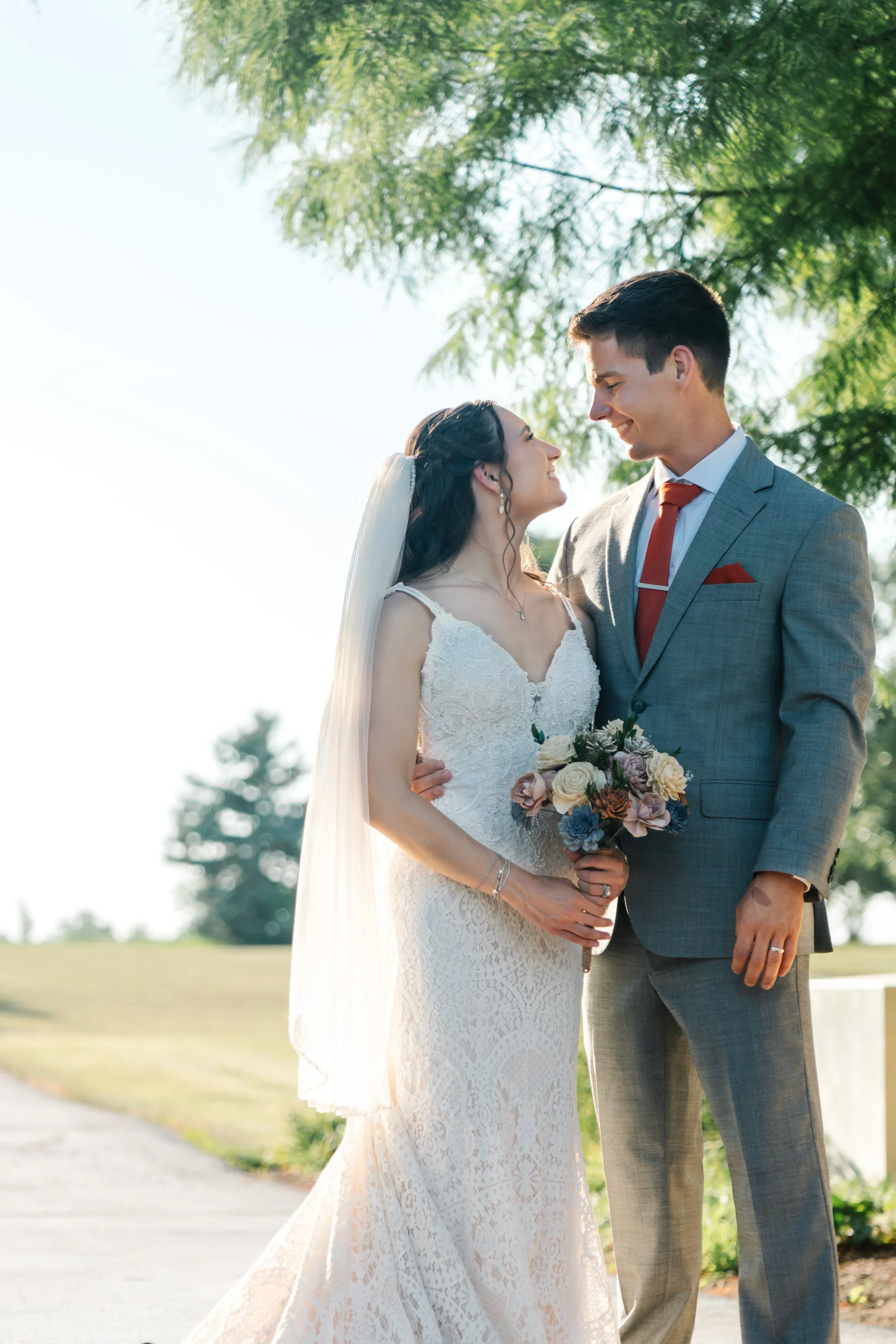 A bride and groom standing close together outdoors, smiling and gazing into each other's eyes, with a tree and bright sky in the background. The bride wears a lace wedding dress and holds a bouquet, the groom wears a gray suit with a red tie.