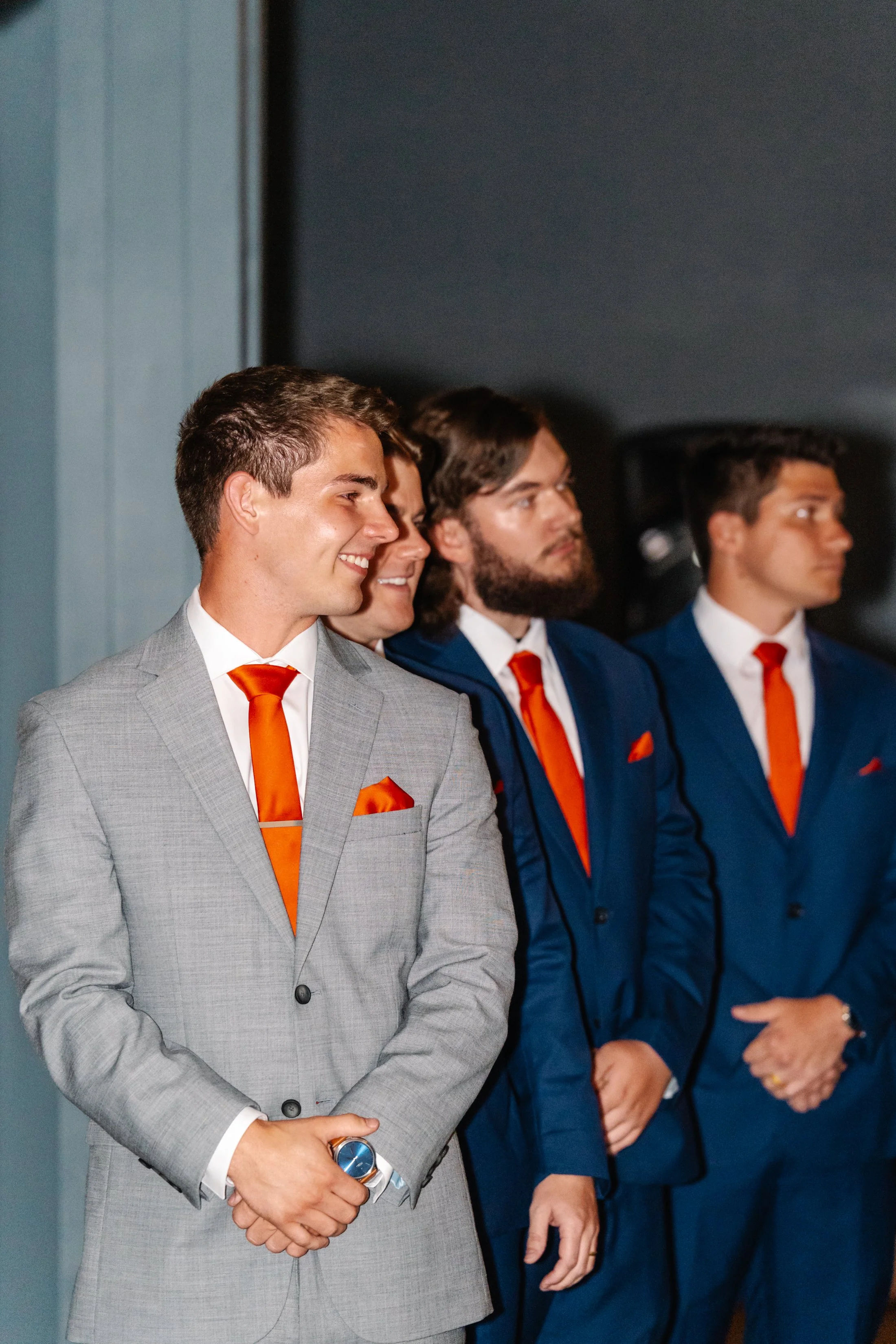 Group of four men dressed in suits with orange ties and pocket squares, standing indoors, at a formal event.