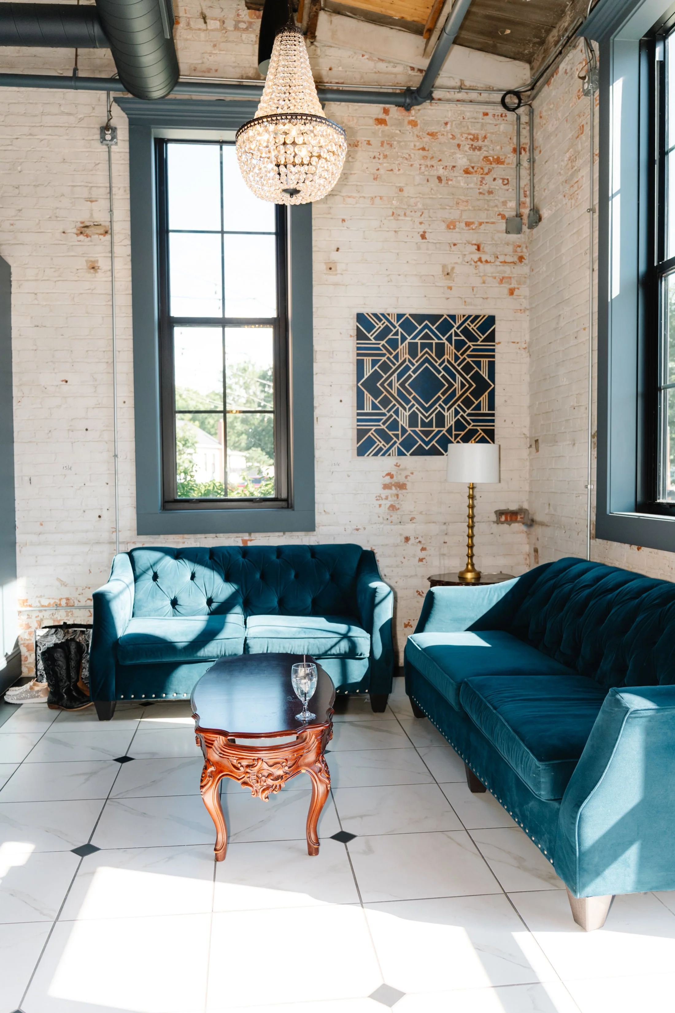 Living room with two teal velvet sofas, a wooden coffee table with a glass of water, a white and gold table lamp, a blue geometric wall art, large black-framed windows, a brick wall, and a chandelier hanging from an industrial ceiling.