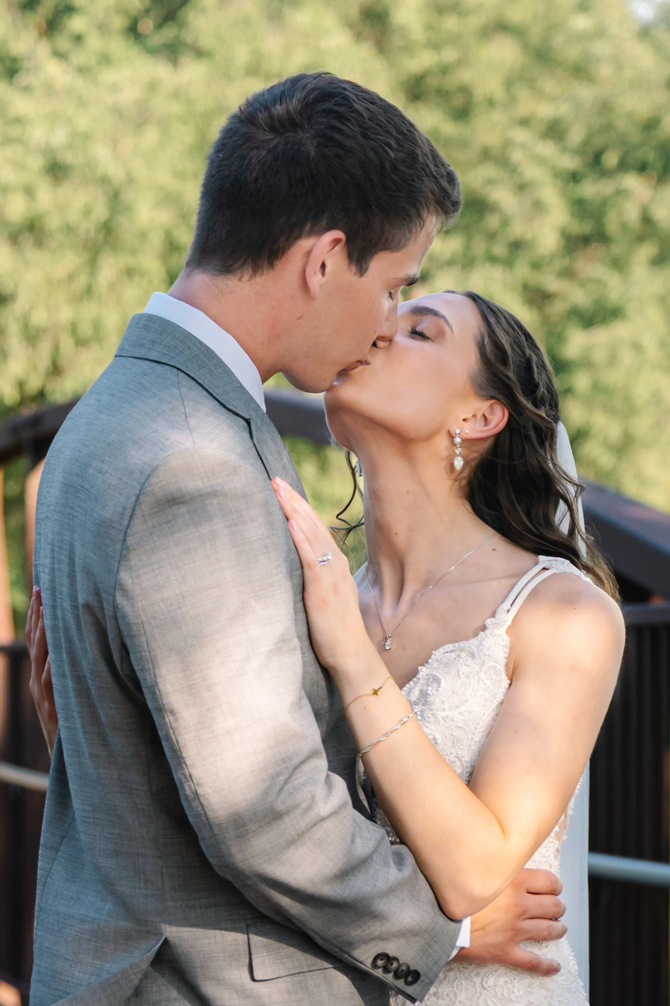A newlywed couple sharing a kiss outdoors, the groom in a gray suit and the bride in a lace wedding dress, against a background of green trees.