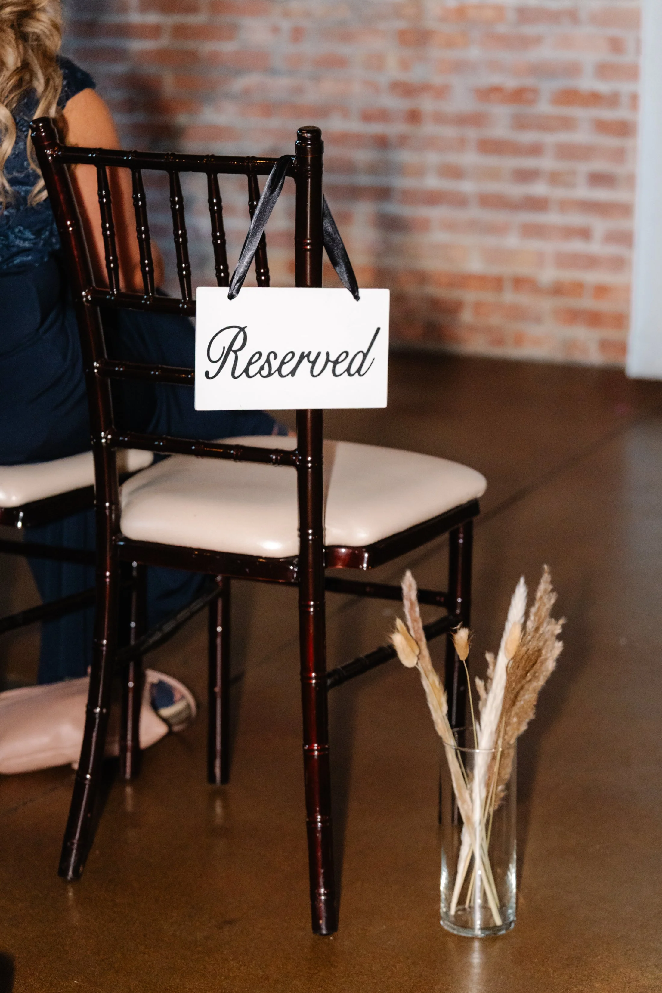 A reserved sign hanging on a dark brown chair with a white cushion at a table in a venue with a brick wall in the background. A glass with dried ornamental grasses is on the table.