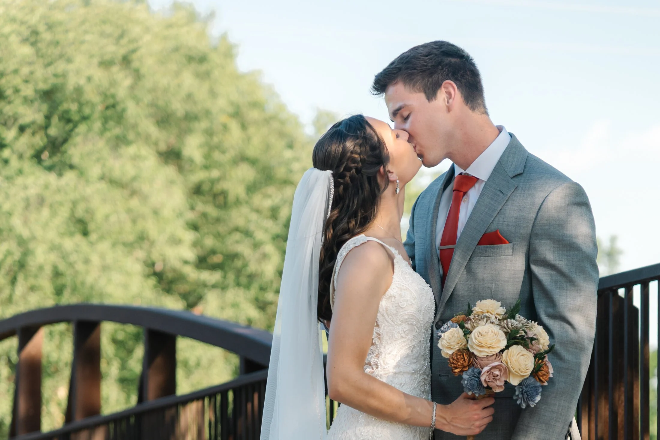 A bride and groom kissing outdoors on a bridge, with green trees in the background. The bride is holding a bouquet of flowers.
