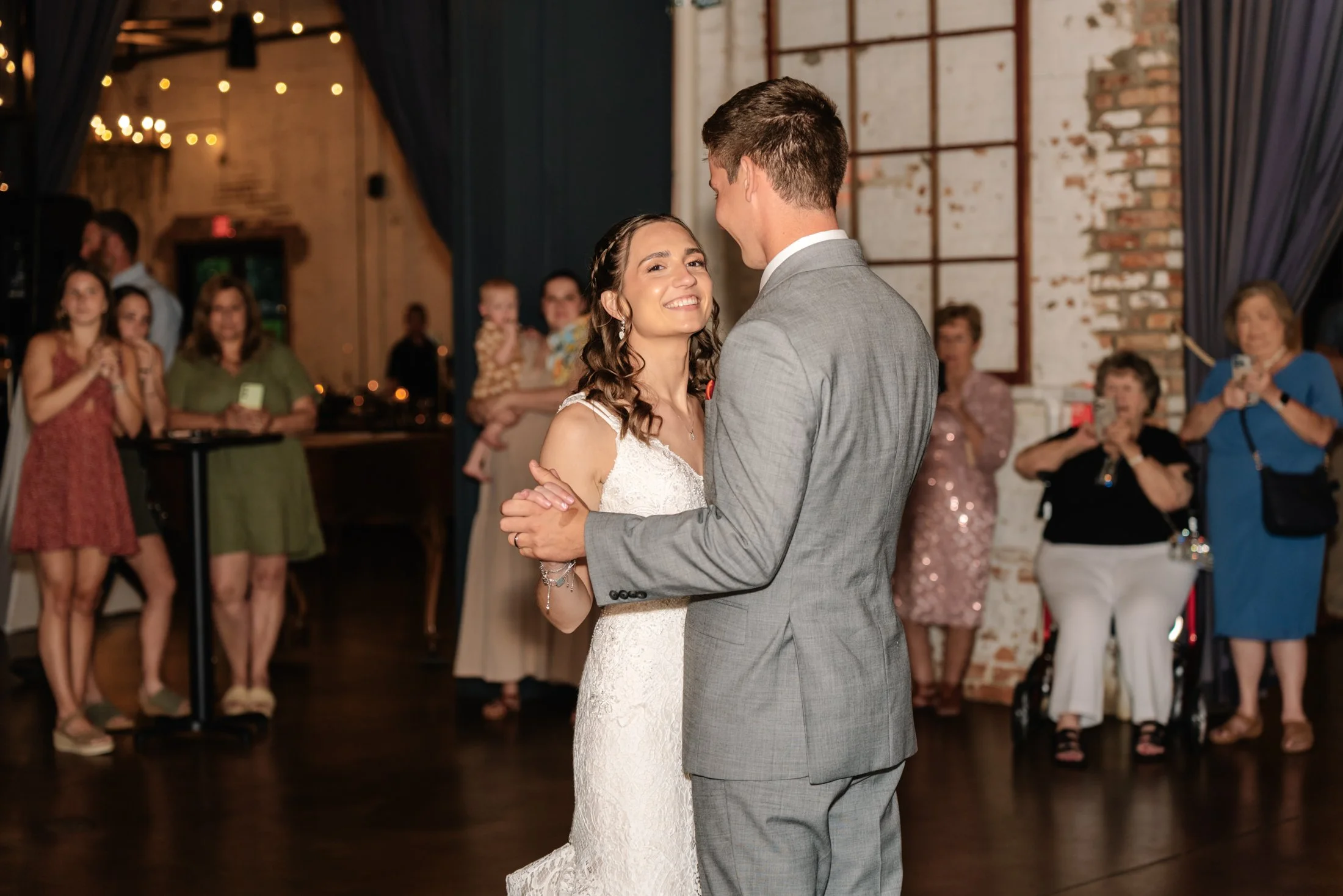 A bride and groom are dancing together at their wedding reception, smiling and looking at each other, while family and friends watch in the background.