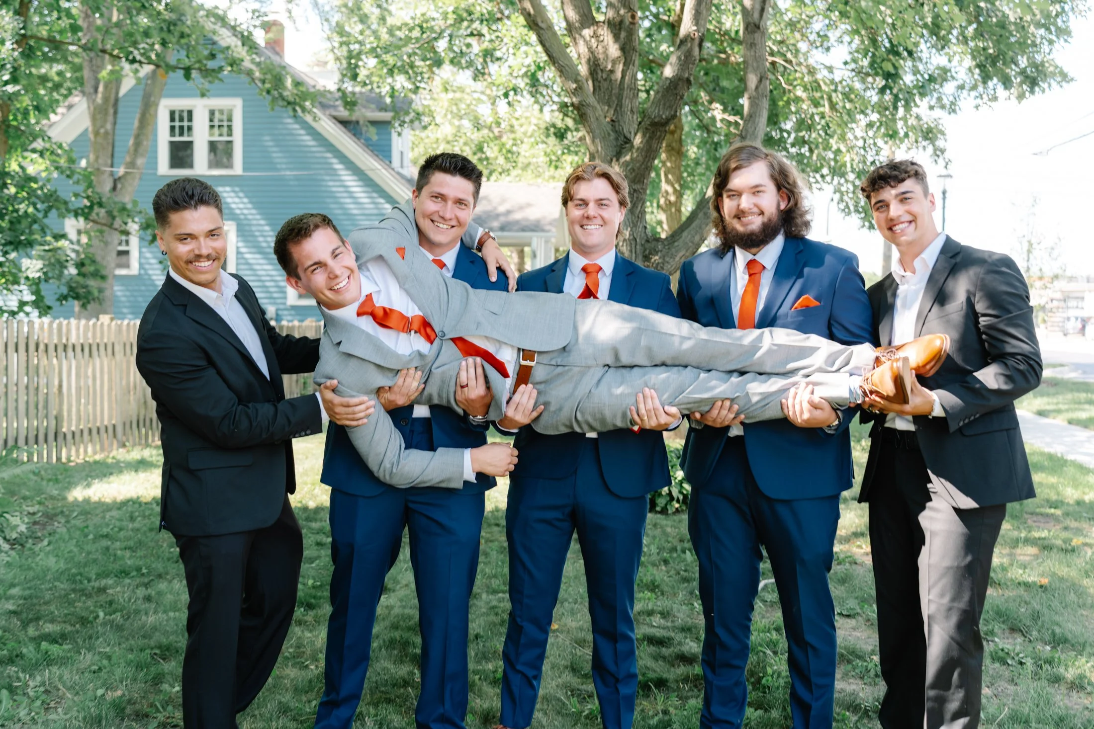 A group of six young men dressed in suits, standing outdoors in front of a house and trees. Five men are holding a man horizontally across their arms, and the man being held is wearing a light gray suit with a red tie.