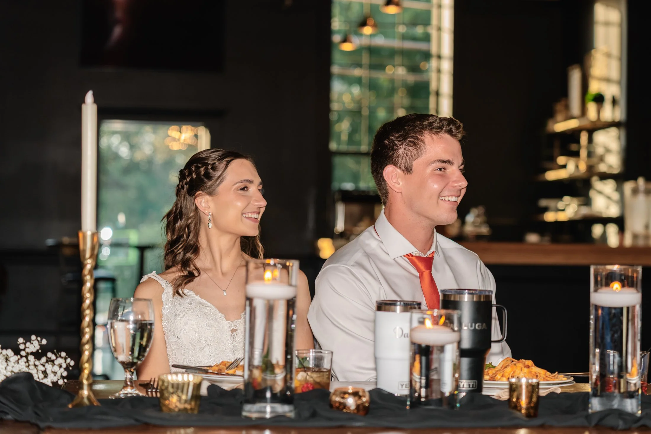 A smiling couple, a woman in a white dress with lace details and a man in a white shirt with an orange tie, sitting at a dinner table with candles and candles on the table, in a warmly lit indoor setting.