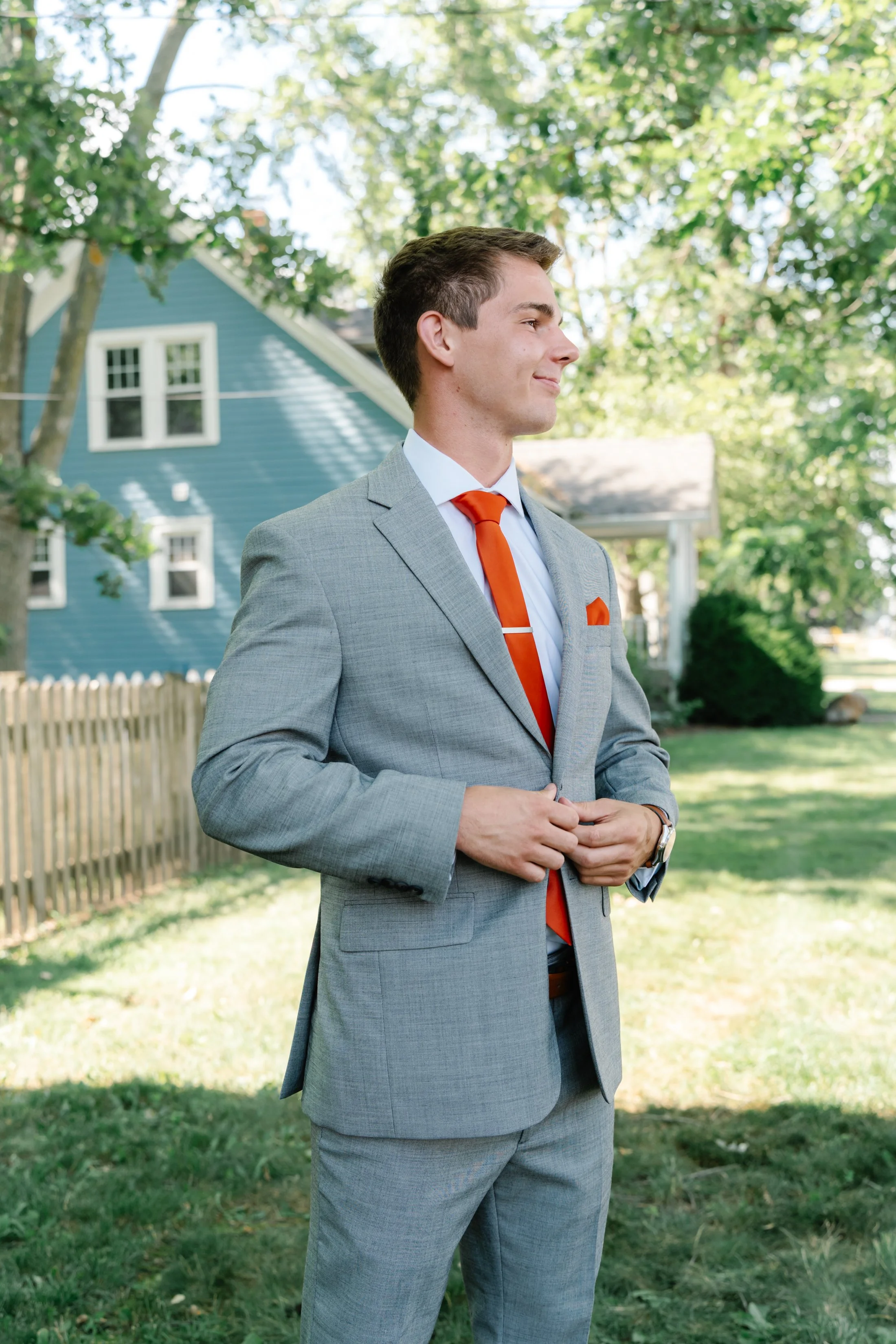 Young man in a gray suit with an orange tie standing outdoors in a backyard with green trees and a blue house in the background, smiling with his hands clasped.