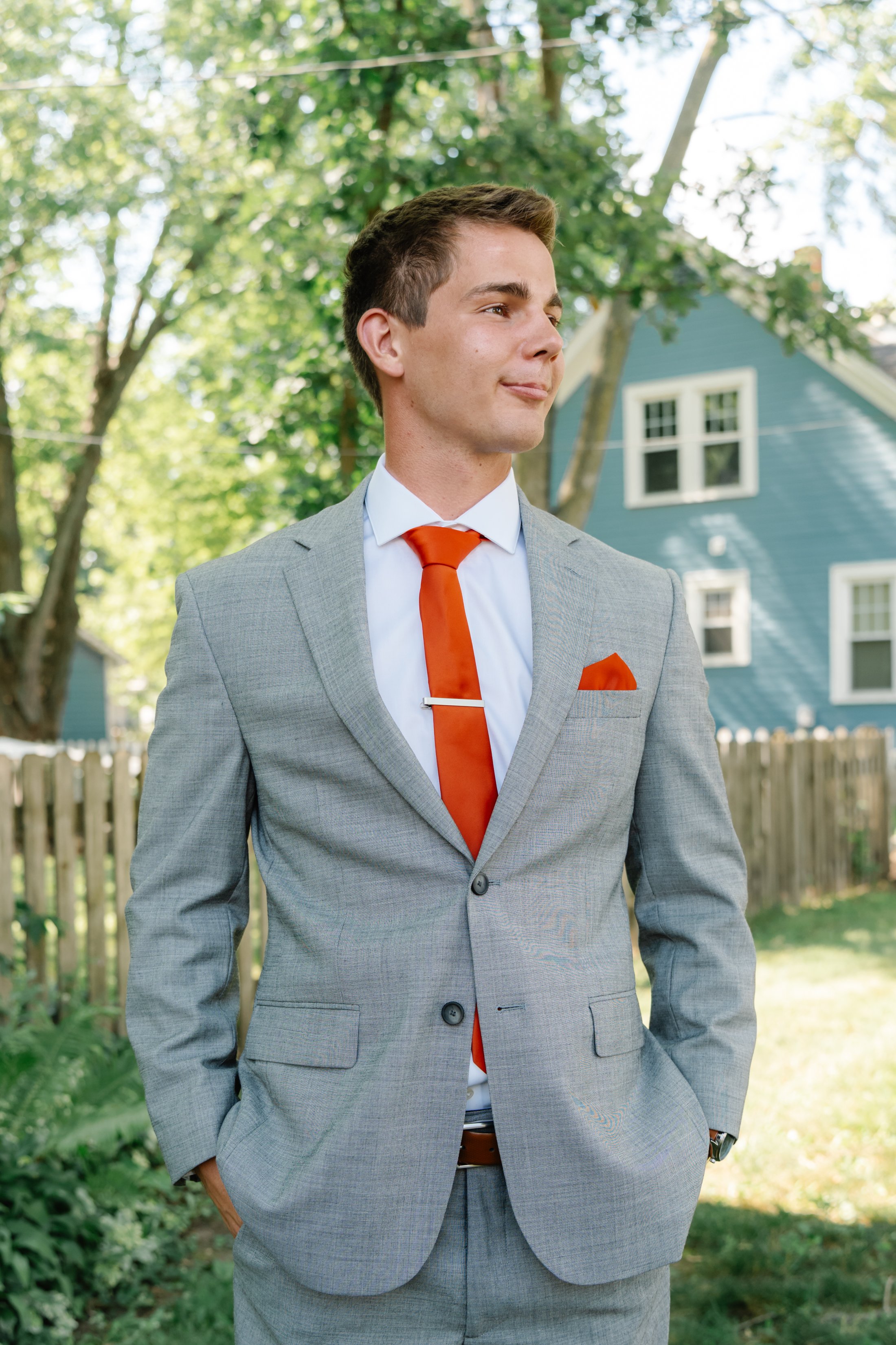 A young man in a gray suit with an orange tie and pocket square stands outdoors in front of a blue house and trees.