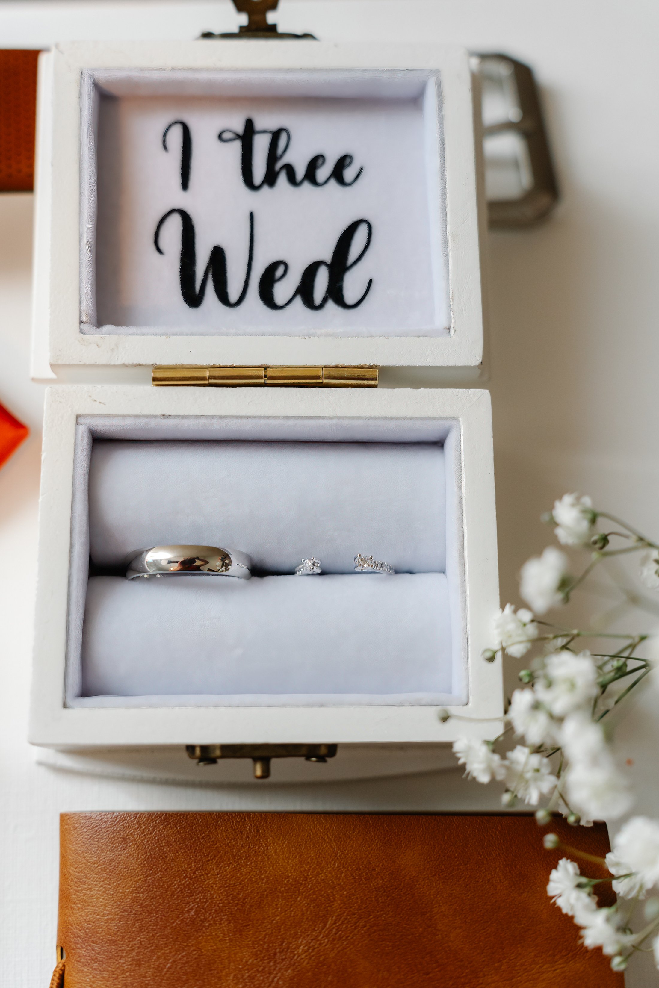 An open white jewelry box with the words 'I thee Wed' inside the lid, showing a wedding band and earrings, with white flowers and a brown leather item partially visible around it.