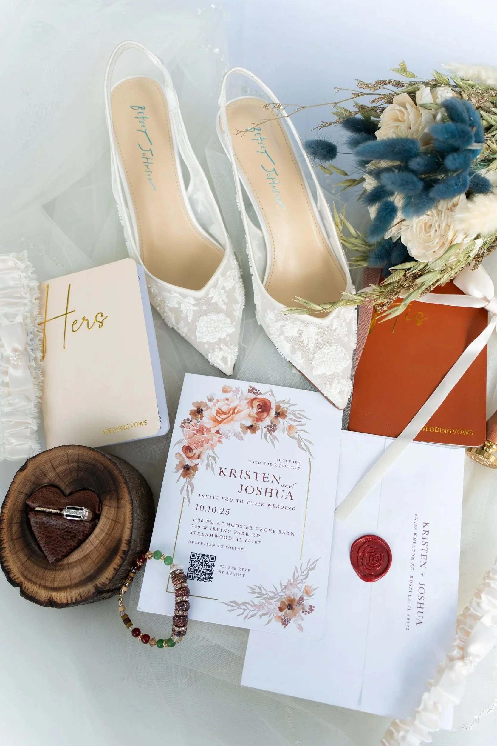A pair of white bridal shoes with lace details, wedding invitations for Kristen and Joshua, a red bouquet with white and blue flowers, a small wooden slab with jewelry, and an envelope with a red wax seal.