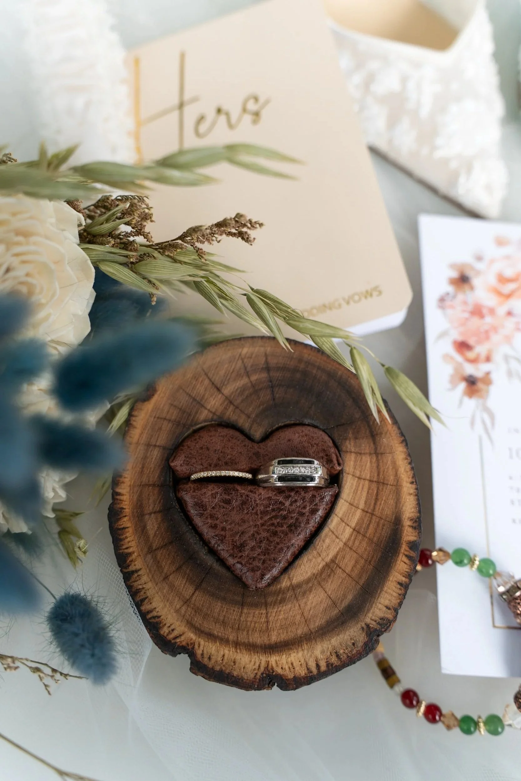 Wedding rings placed on a wooden heart-shaped dish inside a log slice, surrounded by flowers and wedding invitations.