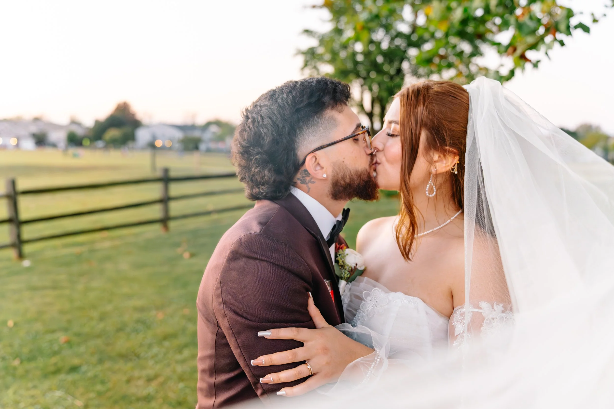 A newlywed couple sharing a kiss outdoors in a park, with a black fence and houses in the background during golden hour.