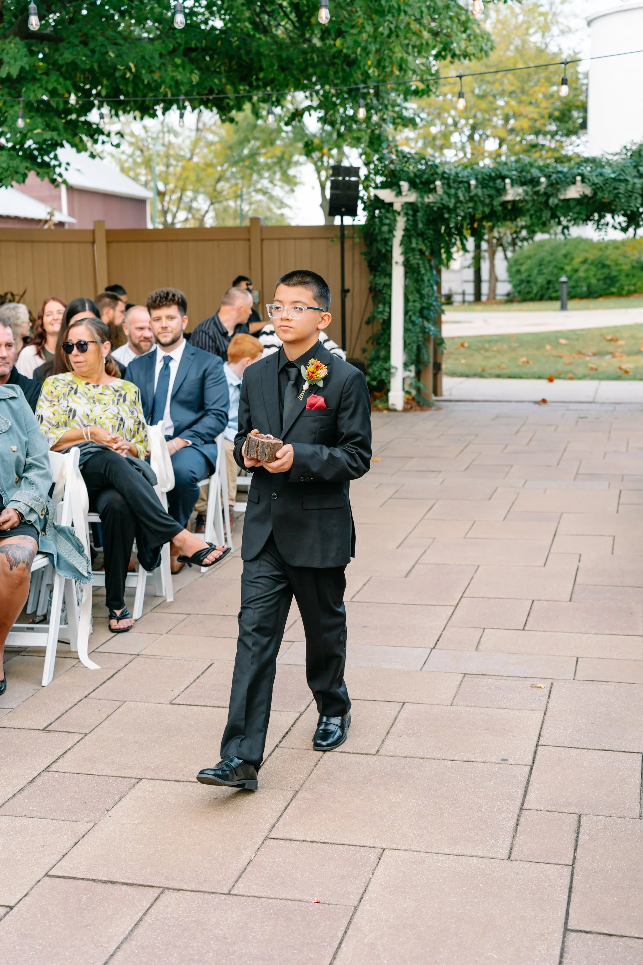 A young man in a black suit with a boutonniere, holding a small box, walking in an outdoor setting during a formal event, with seated guests watching.