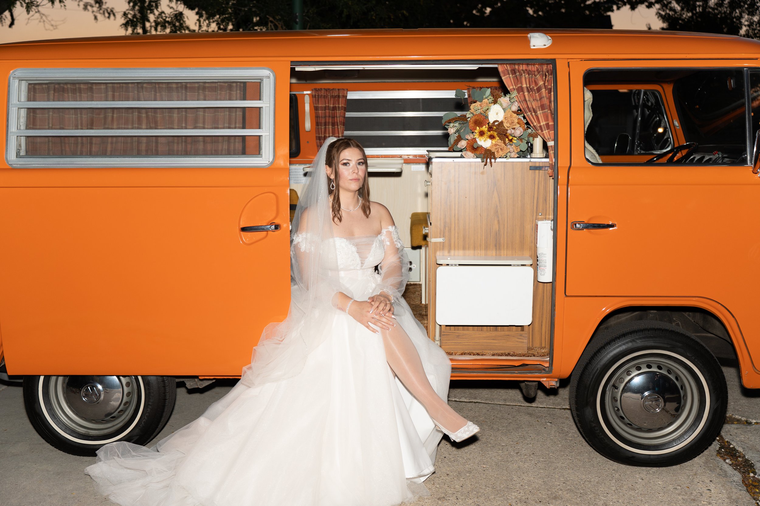 A woman in a white wedding dress and veil sitting on the step of an orange vintage camper van, with a bouquet of flowers inside and curtains in the background.