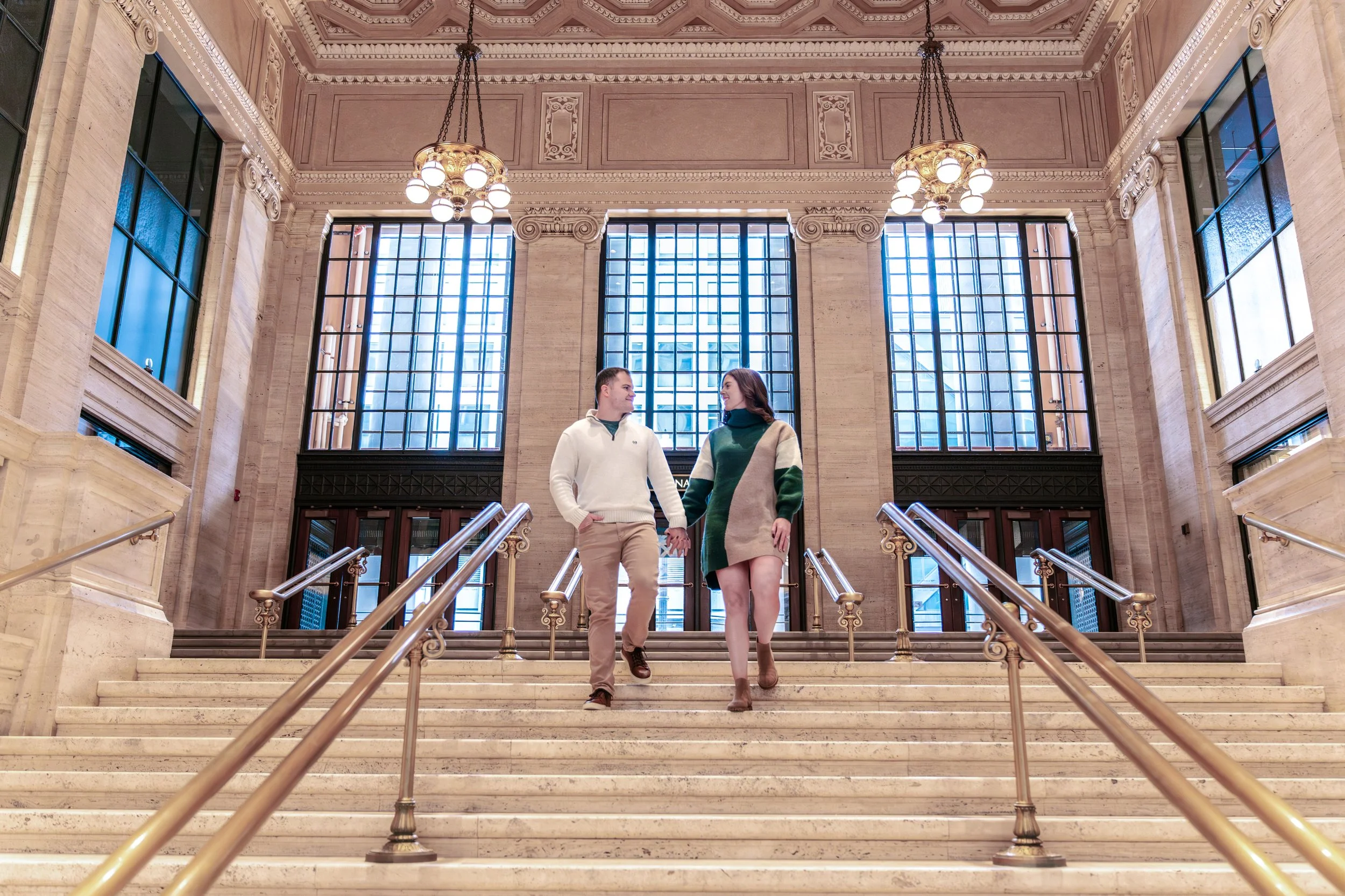 A young couple walking together hand-in-hand down the marble stairs inside a grand building with large windows and ornate chandeliers.
