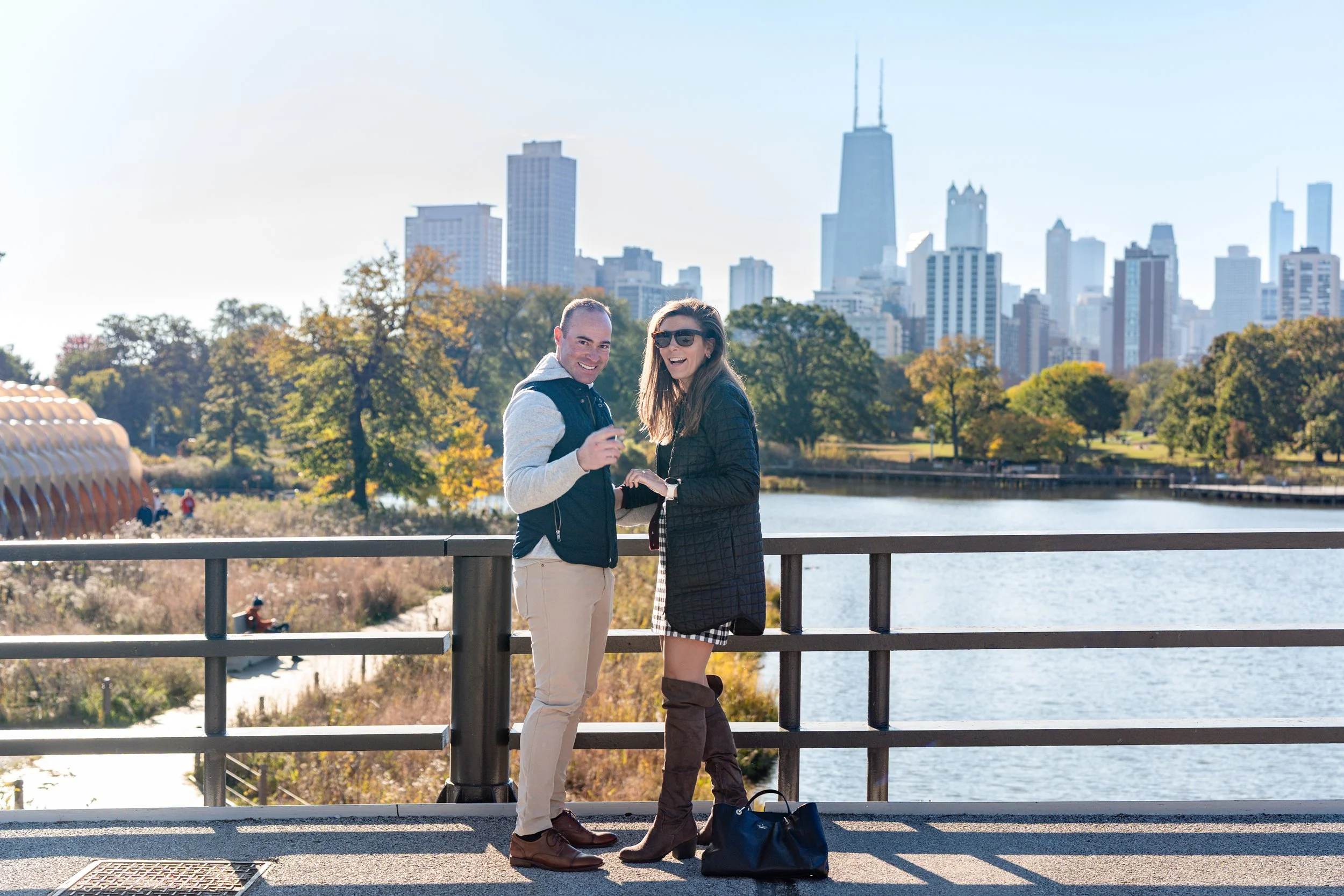 A man and woman standing on a bridge by a lake in a city park with skyscrapers in the background, smiling and enjoying the sunny day.