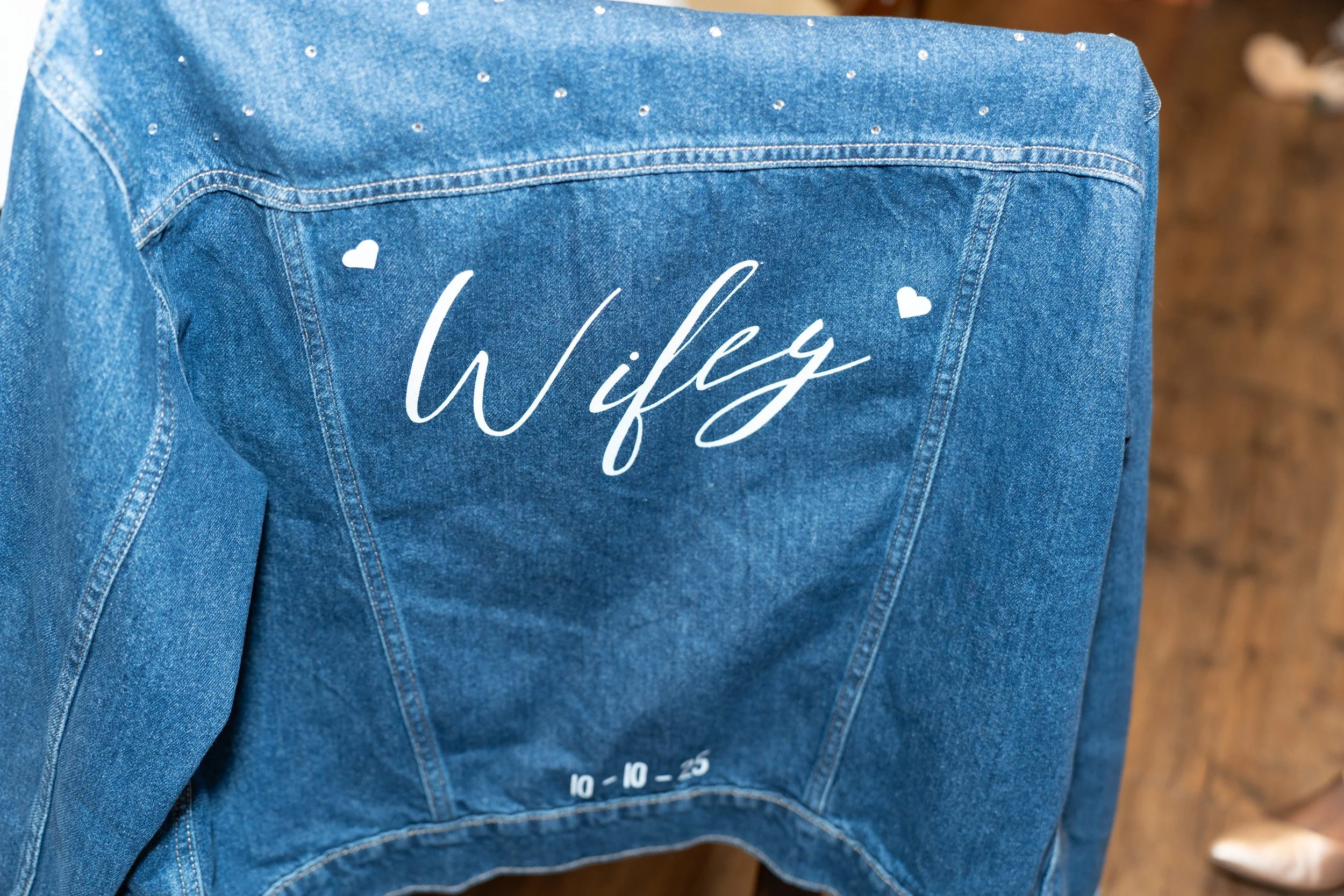Blue denim jacket with white handwritten text 'Wifey' and small white hearts above and below, along with wedding date '10-10-25' at the bottom.