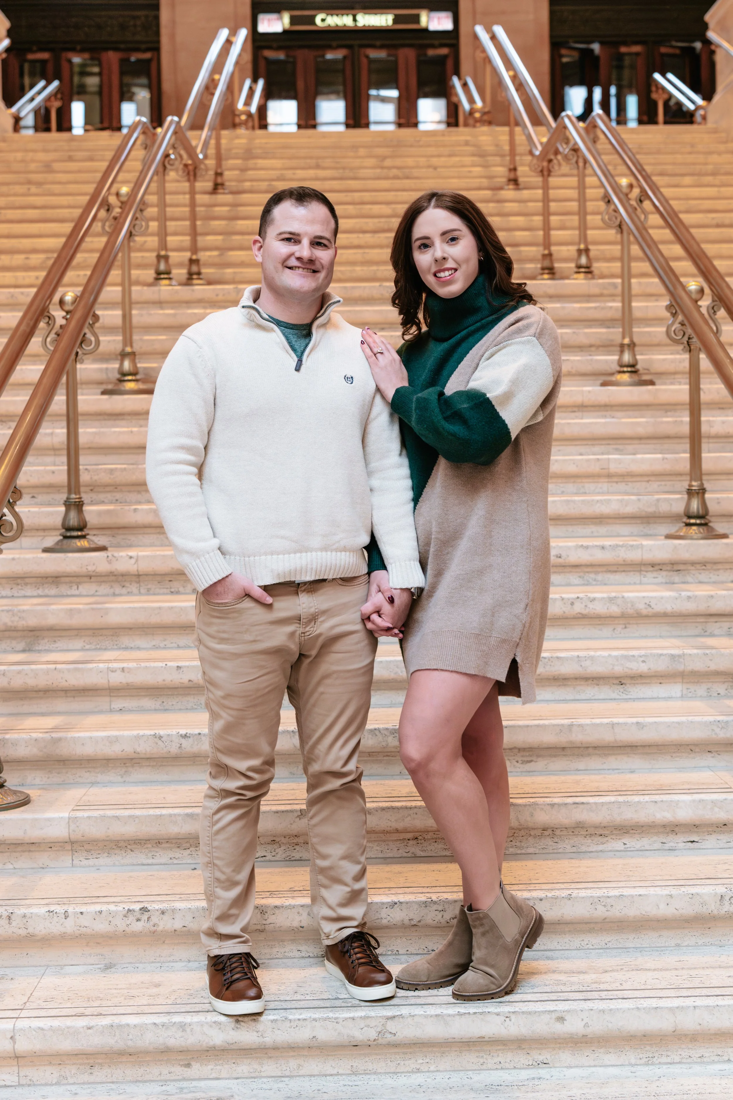 A couple standing on a staircase inside a building, holding hands and smiling at the camera, with a sign that reads 'Calf Street' in the background.