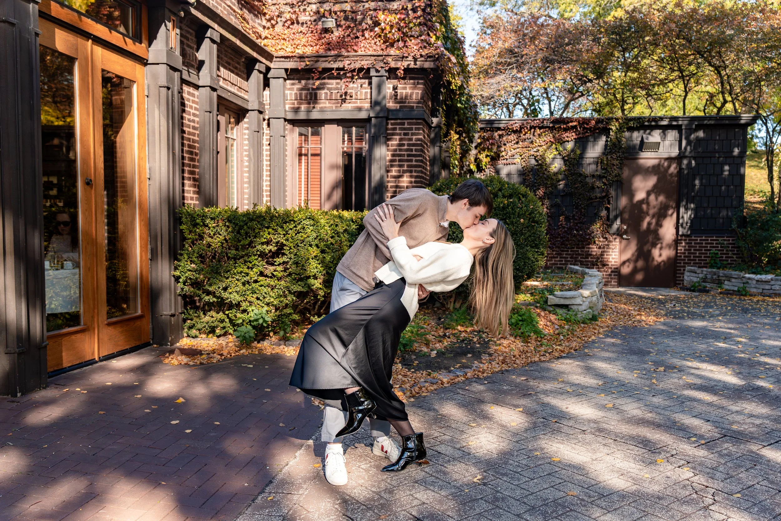 A couple sharing a kiss outside a house with autumn trees and fallen leaves.