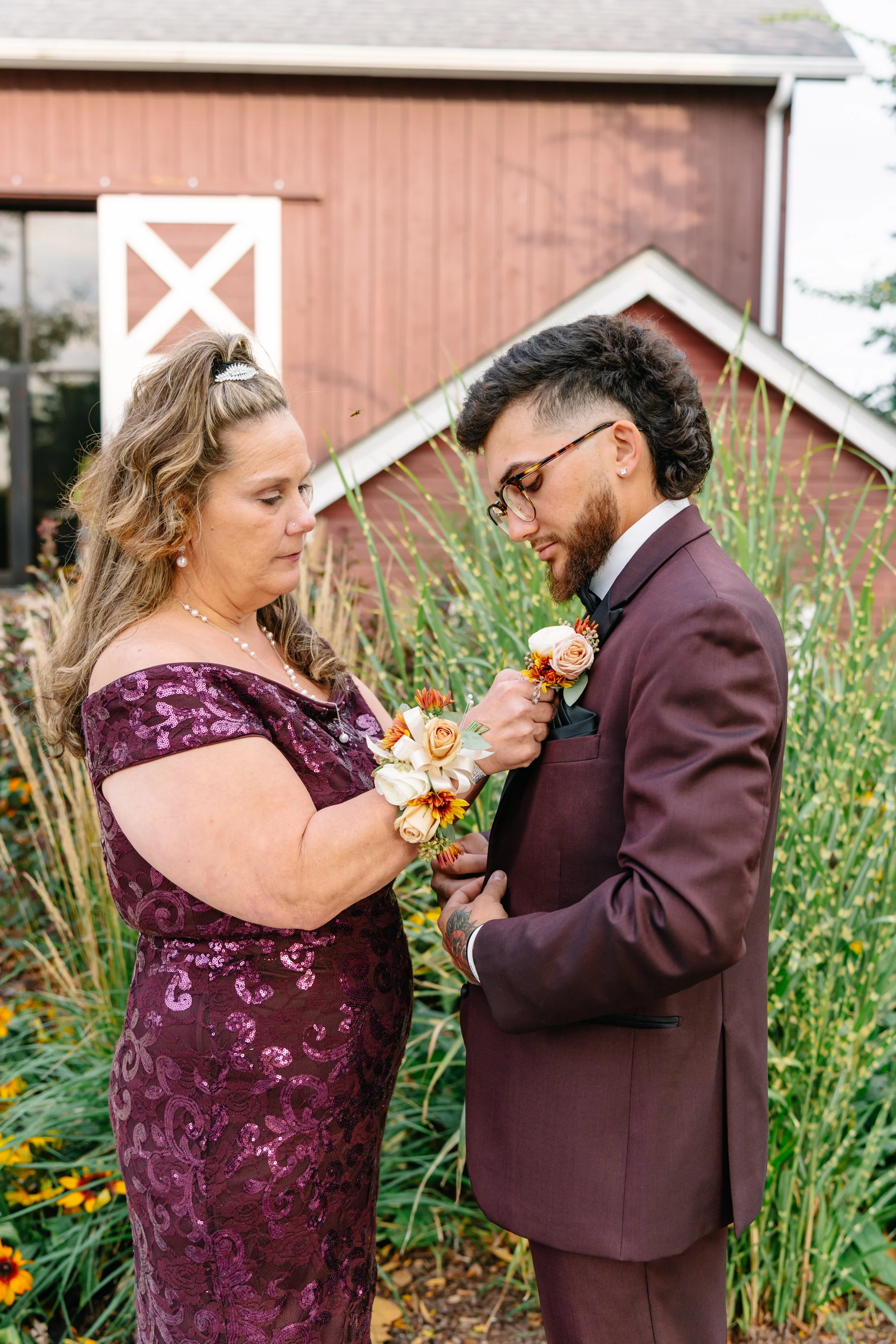 A woman in a purple lace dress pins a boutonniere onto a man in a maroon suit during a wedding ceremony outdoors, with a red barn and tall grass in the background.