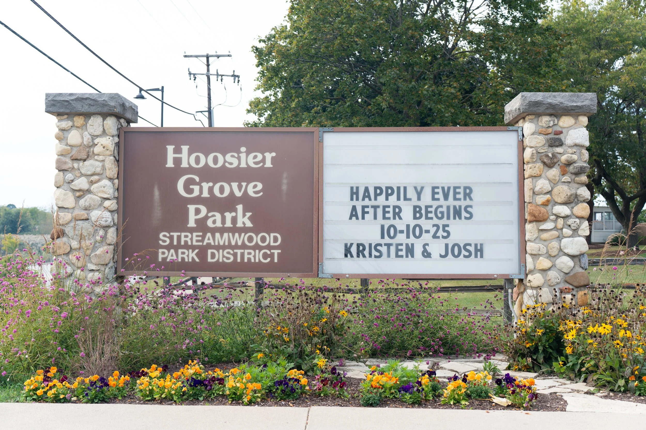 Sign at Hoosier Grove Park in Streamwood Park District, with a message celebrating a wedding anniversary on 10-10-25 from Kristen and Josh, surrounded by colorful flowers and trees.