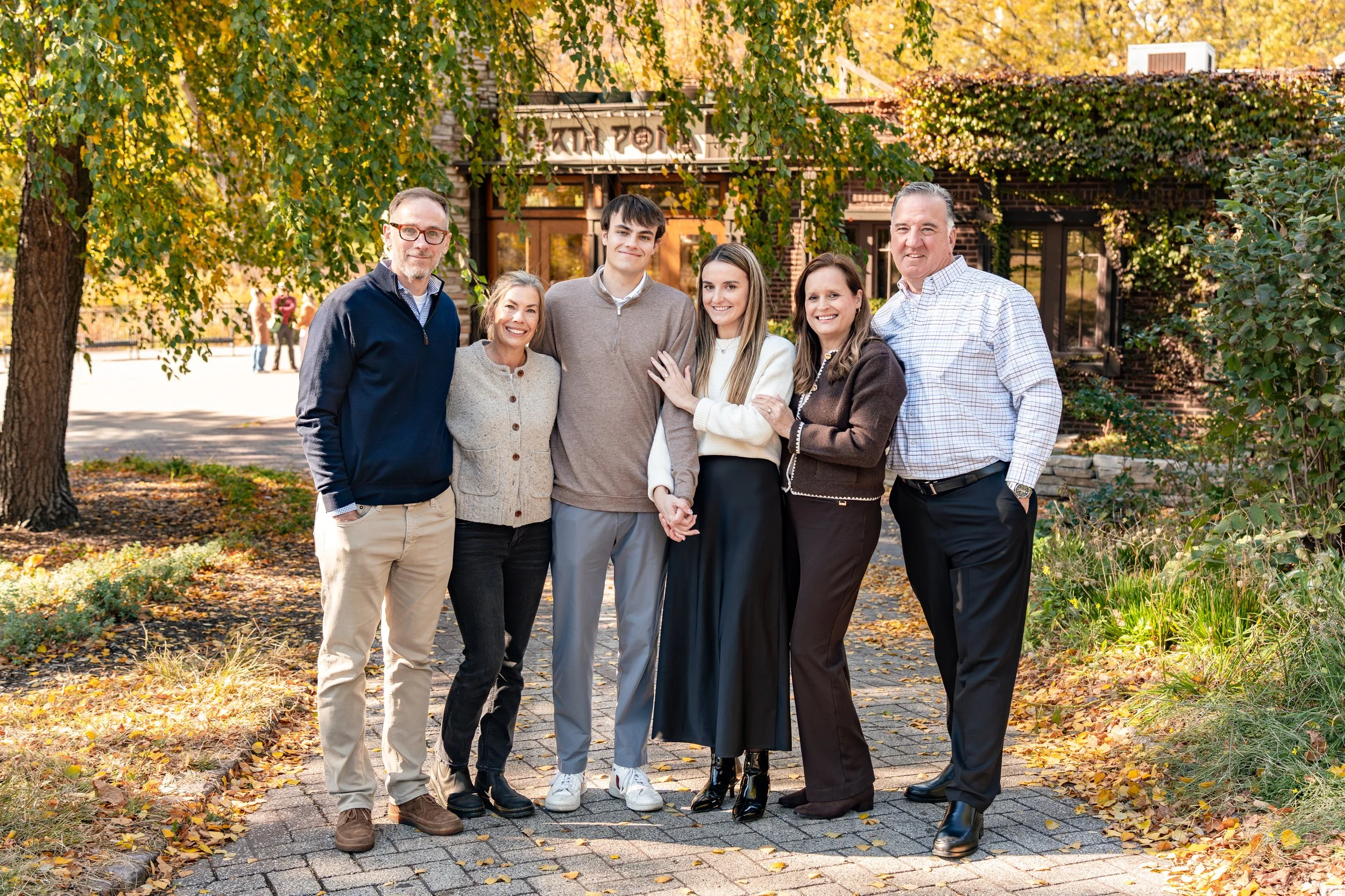 Family group of six people standing outdoors in fall, smiling, in front of a building with trees and fallen leaves.