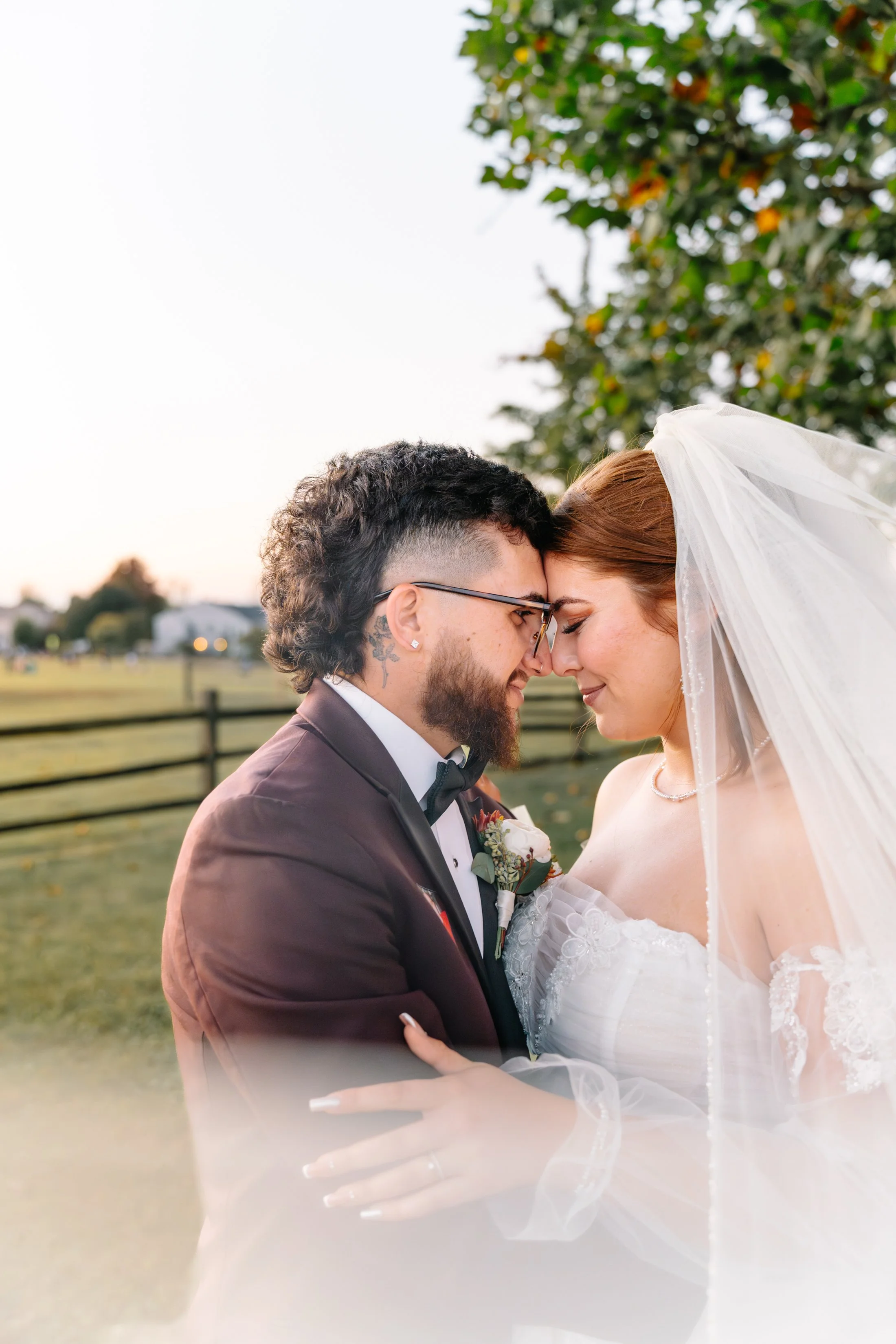A newlywed couple with foreheads touching, eyes closed, outdoors during sunset, with a green tree and fence in the background.