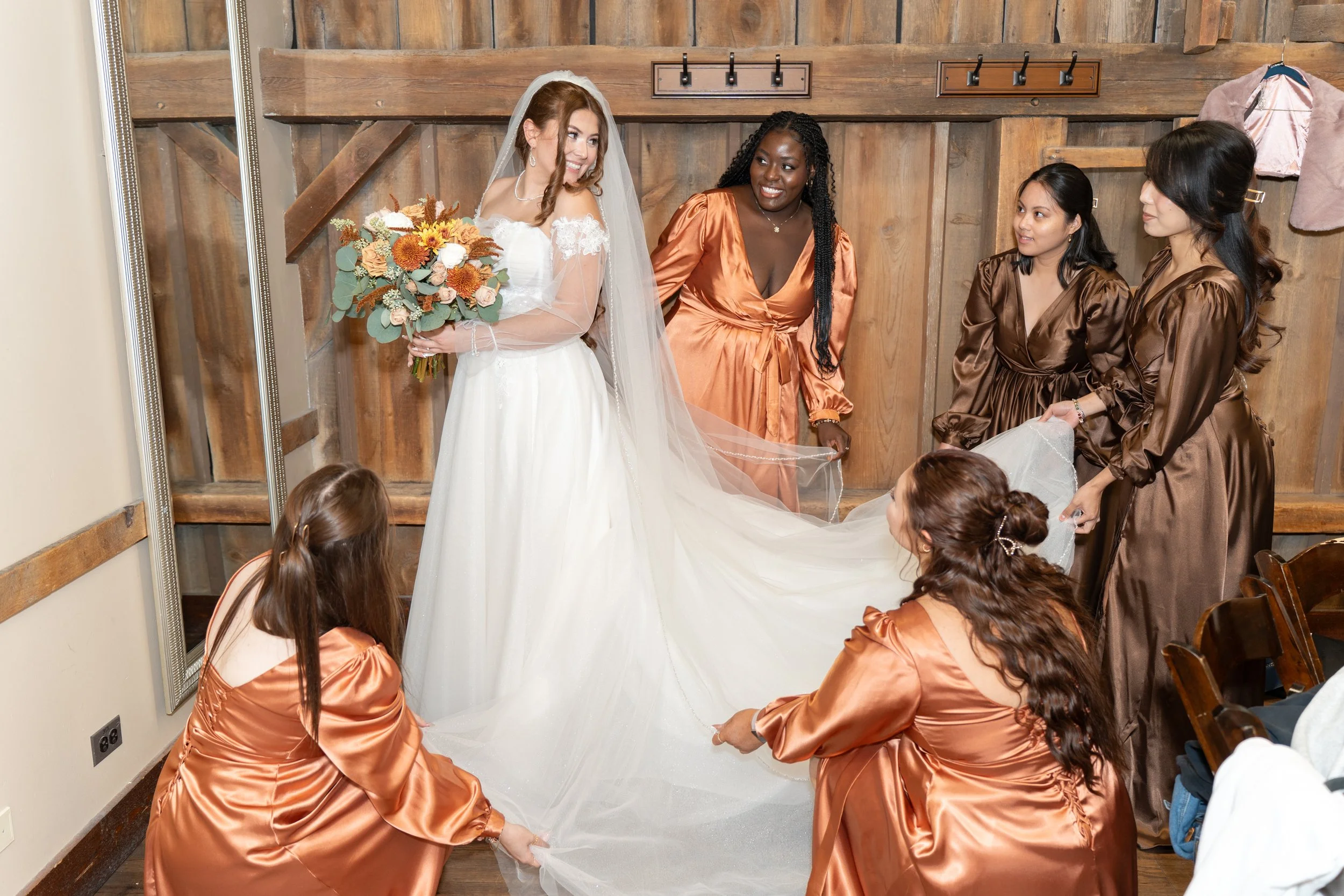 A bride in a white wedding dress and veil standing with a bouquet of flowers, surrounded by bridesmaids in brown satin dresses holding and adjusting her long wedding veil.