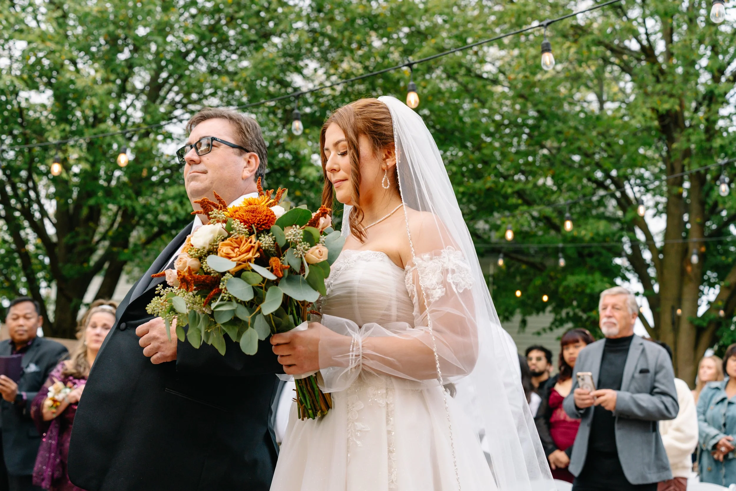 A bride in a white wedding dress holding a bouquet of flowers during an outdoor wedding ceremony, standing beside a man in a black suit with glasses. Several guests are in the background, some using phones, with string lights hanging above and green 