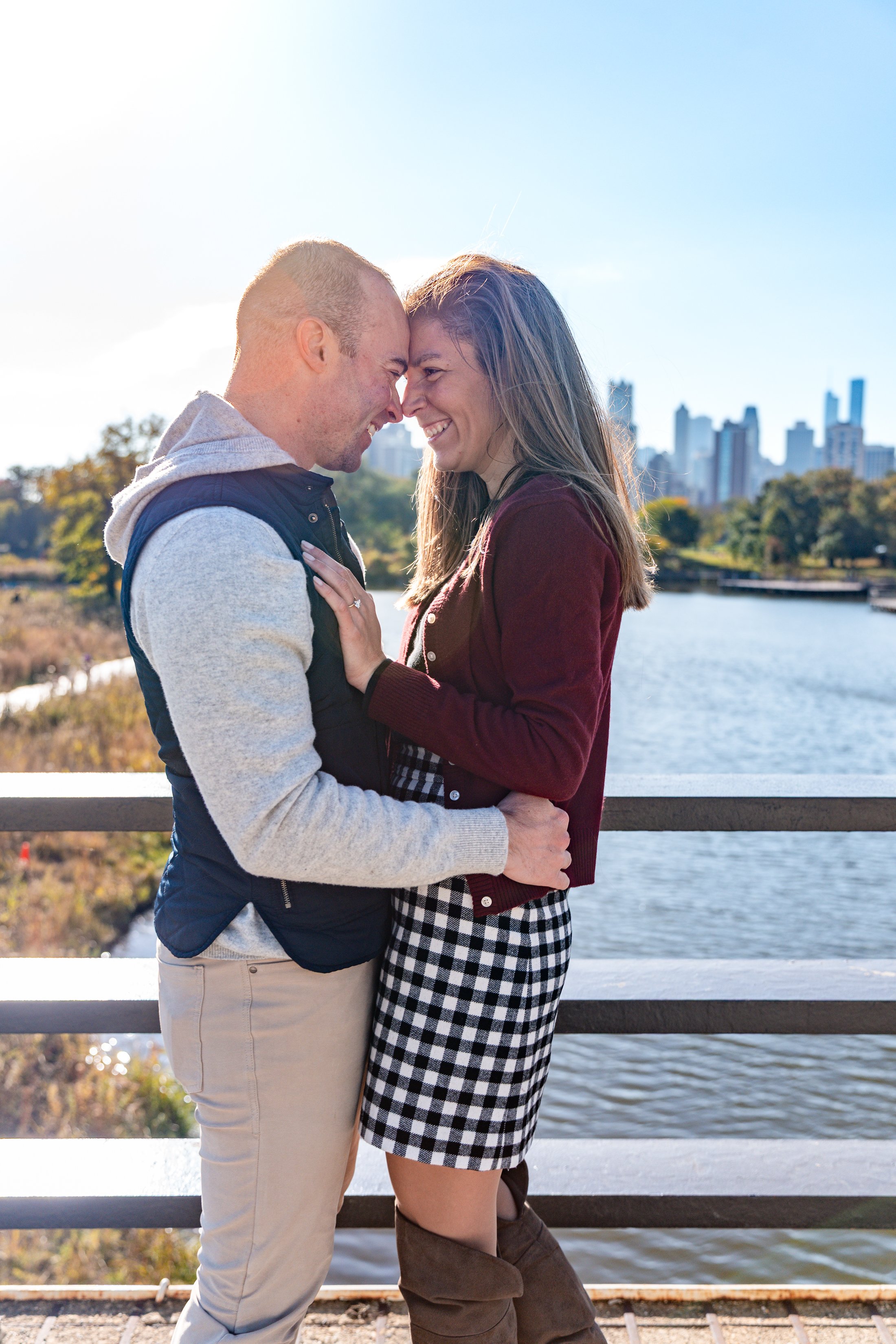 A couple standing close together outside by water, touching foreheads and smiling, with city skyline in the background.