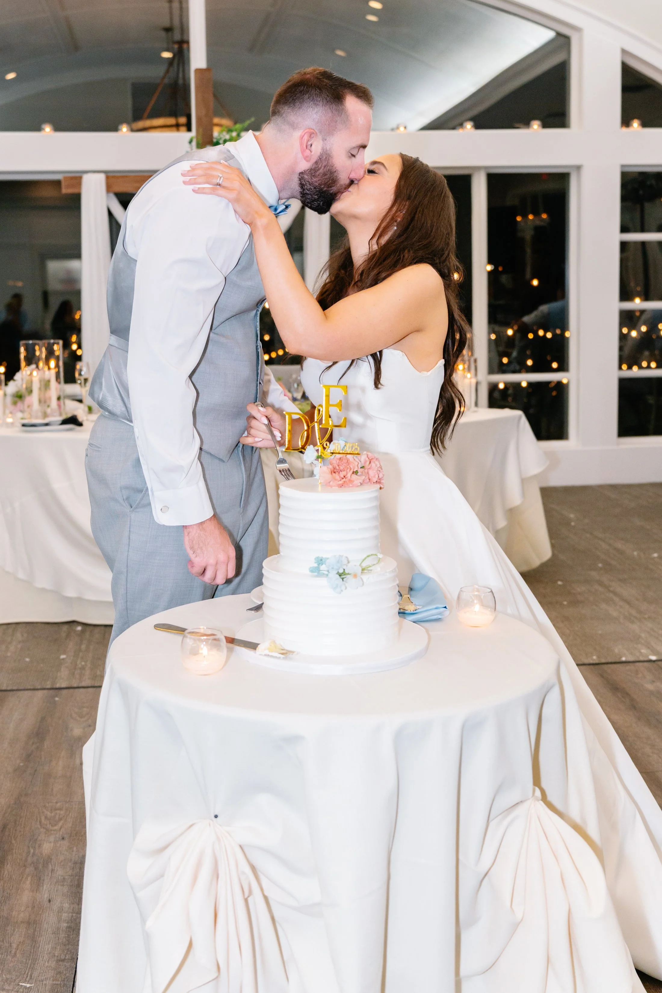 A bride and groom kissing in front of a wedding cake at their reception, with candles and decorated tables in the background.