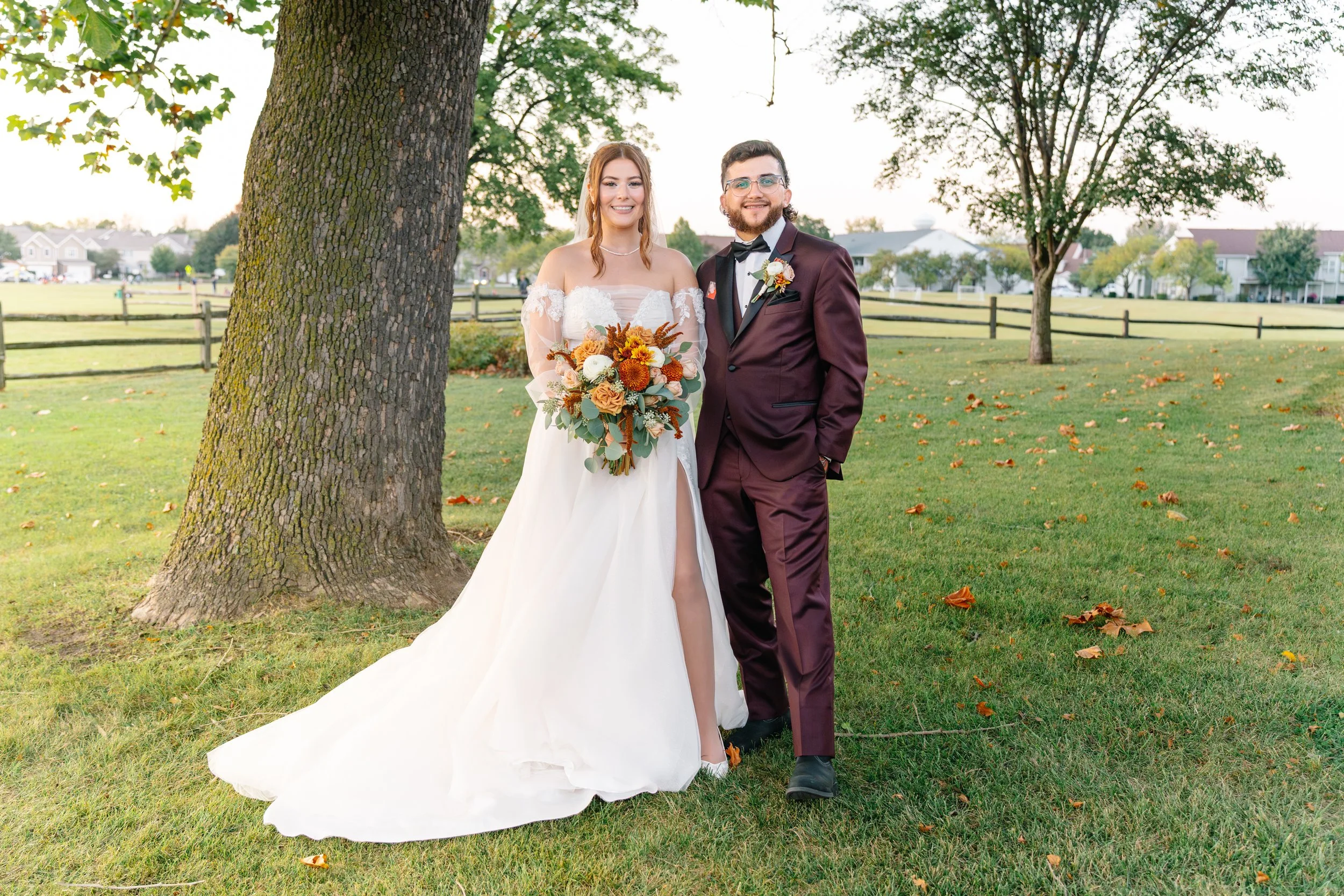A bride in a white wedding dress holding a bouquet of flowers, standing next to a groom in a burgundy suit, outdoor park setting with trees and a fence in the background.