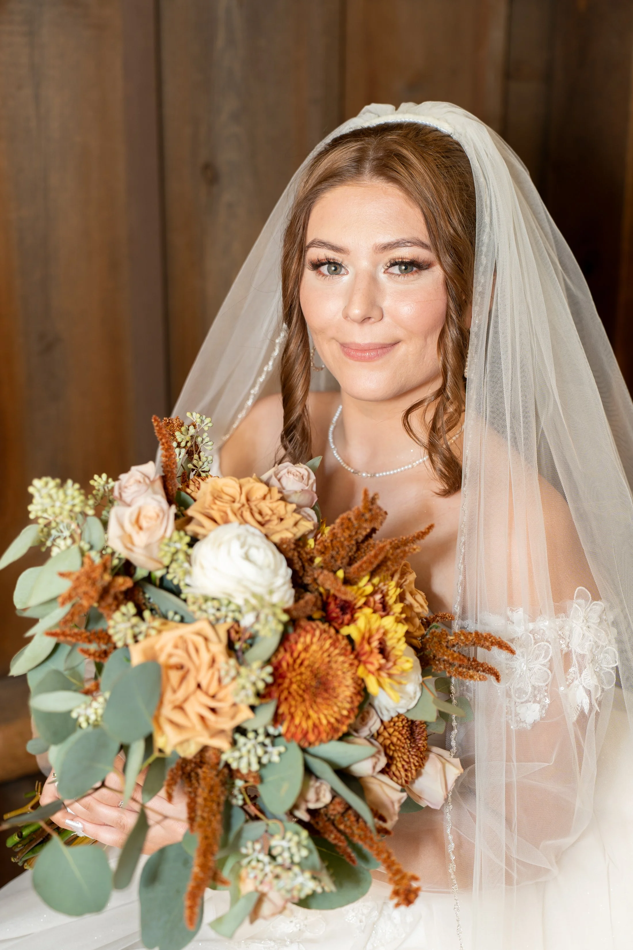 A bride with brown hair wearing a wedding dress and veil, holding a bouquet of flowers, posed in front of a wooden background.