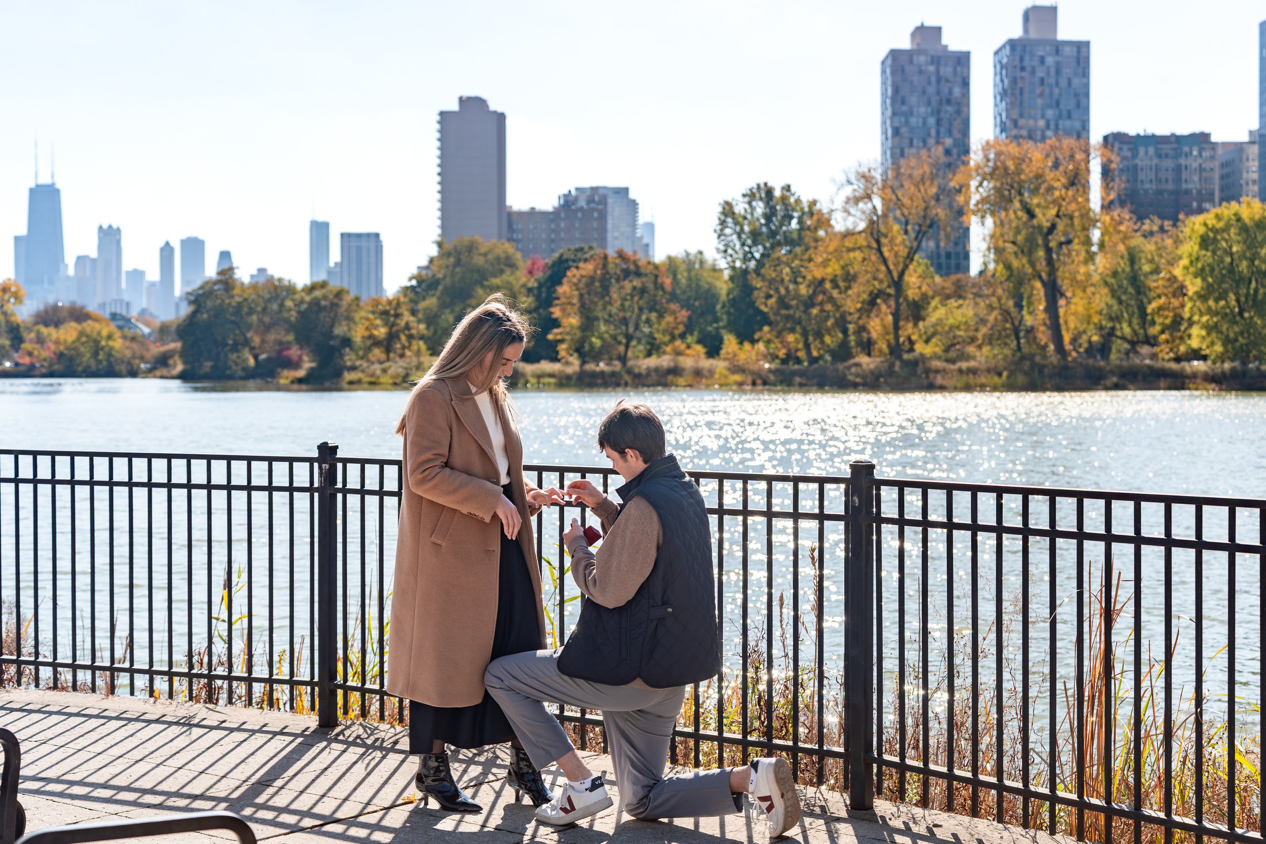 A man proposing to a woman by a lakeside in an urban park during fall, with city skyscrapers in the background.