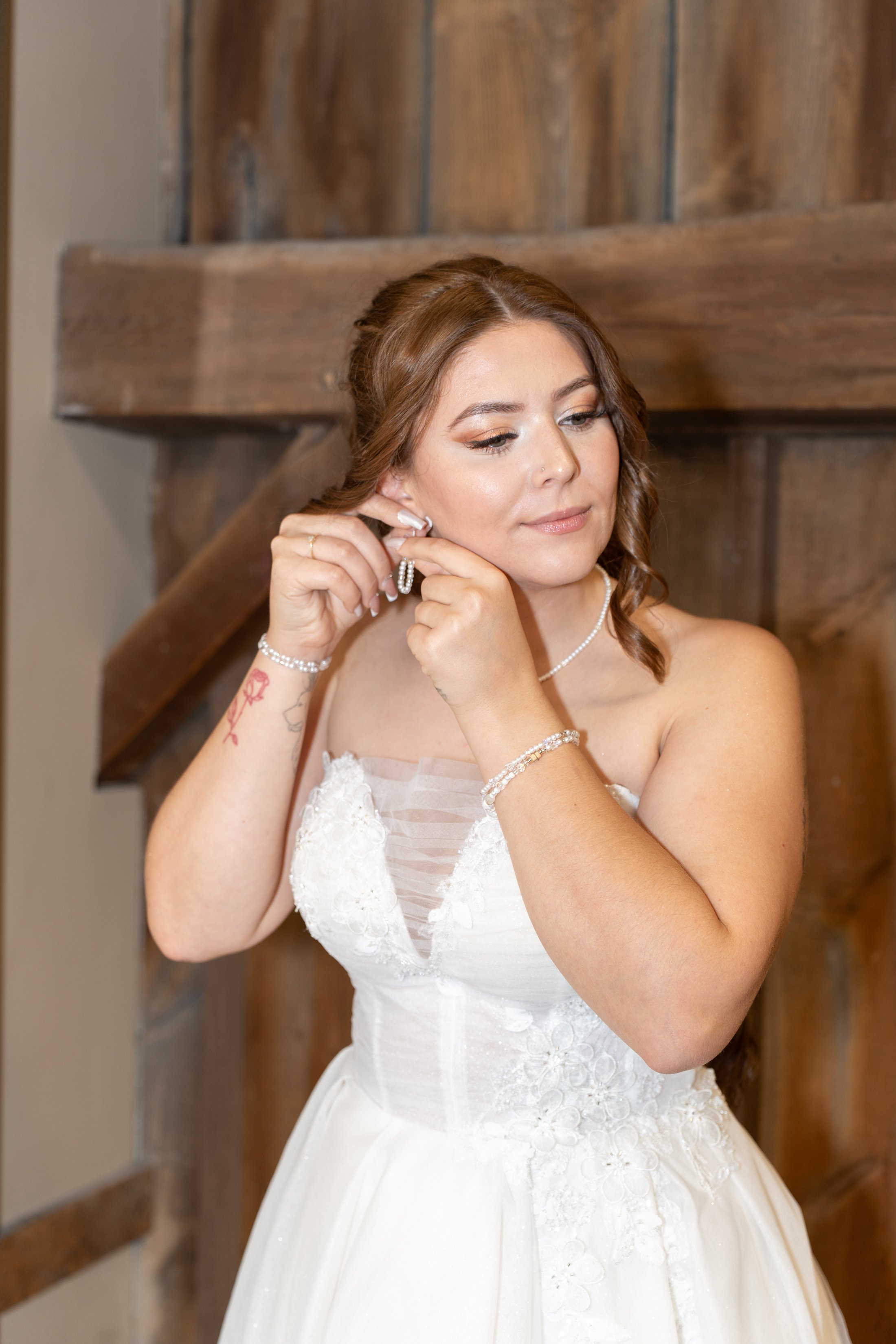 A bride with brown hair in loose curls is putting on earrings. She is wearing a white strapless wedding gown with lace details and a pearl necklace, in front of a wooden wall.